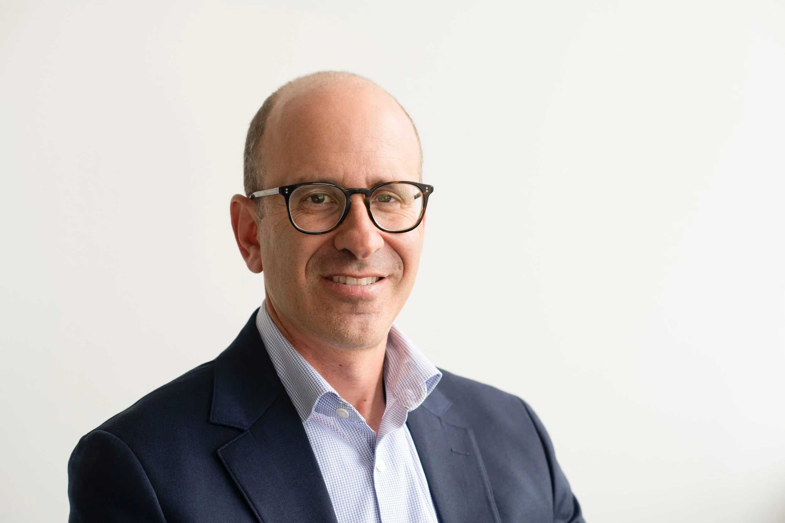 A man with glasses, balding head, wearing a dark suit and a light blue shirt, smiling against a plain white background.