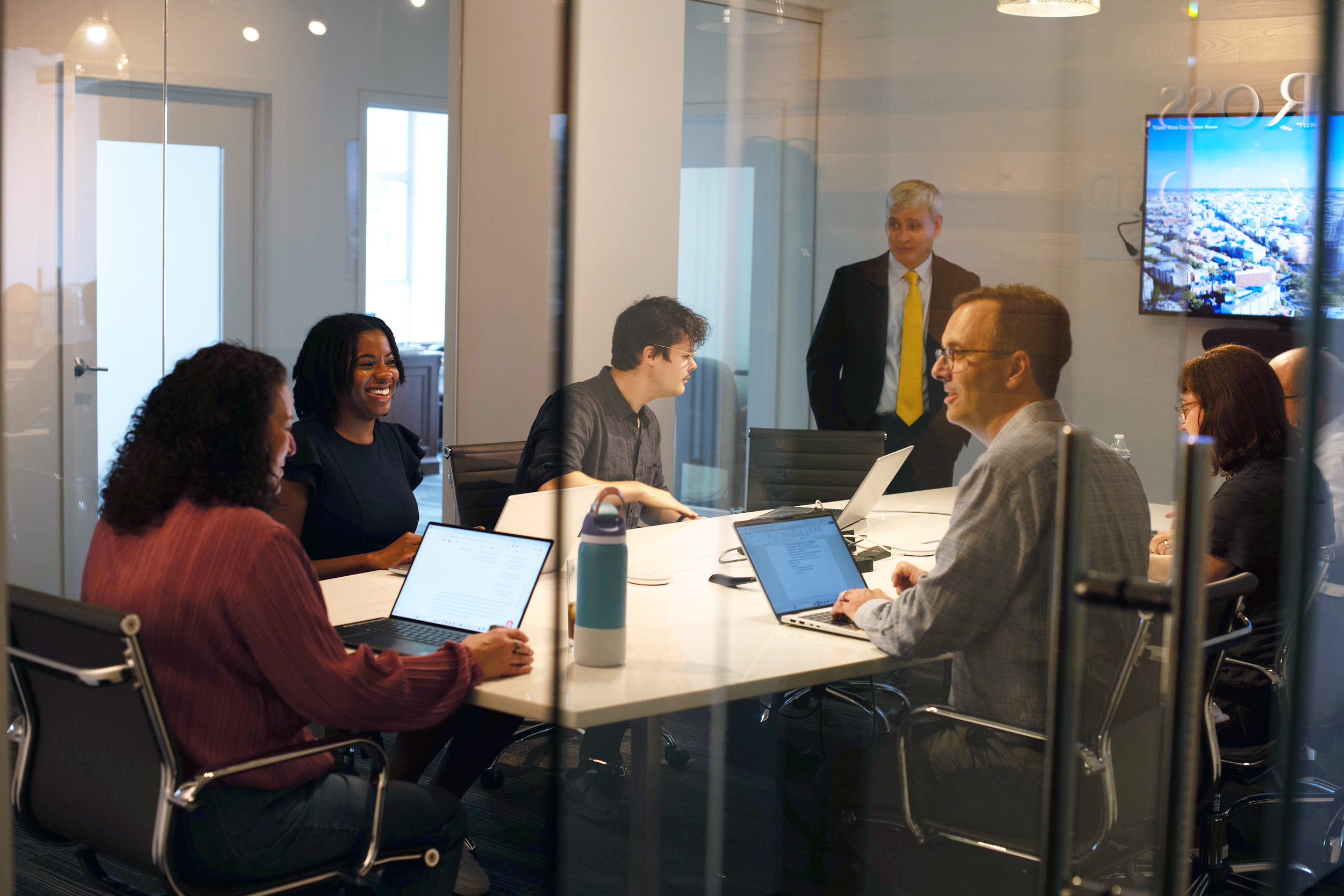 A group of eight diverse professionals engaged in a meeting in a glass-enclosed conference room, with laptops and documents on the table, and a man in a dark suit and yellow tie standing at the head of the table.