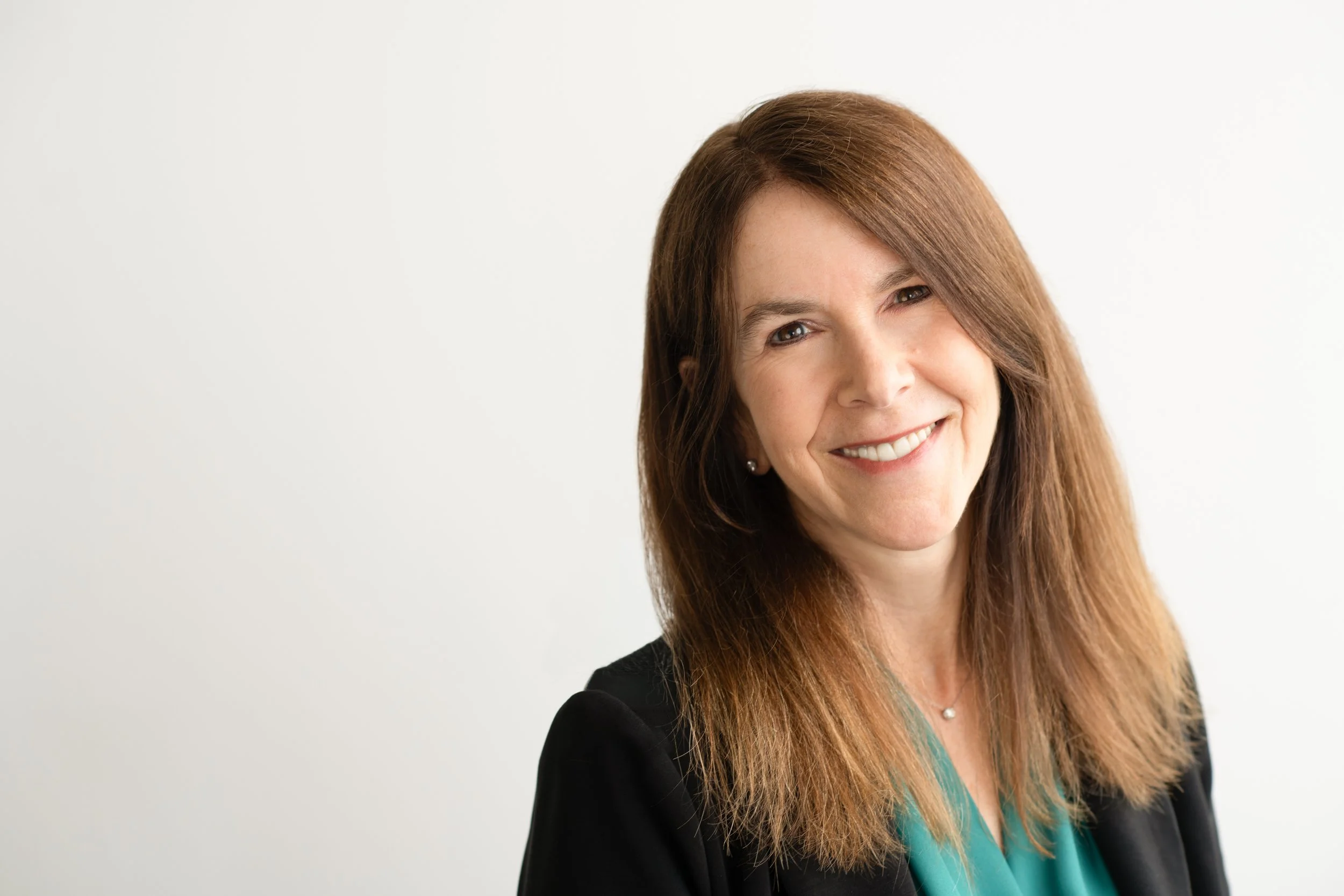 A woman with long, reddish-brown hair smiling, wearing a teal top and a black blazer, against a plain white background.