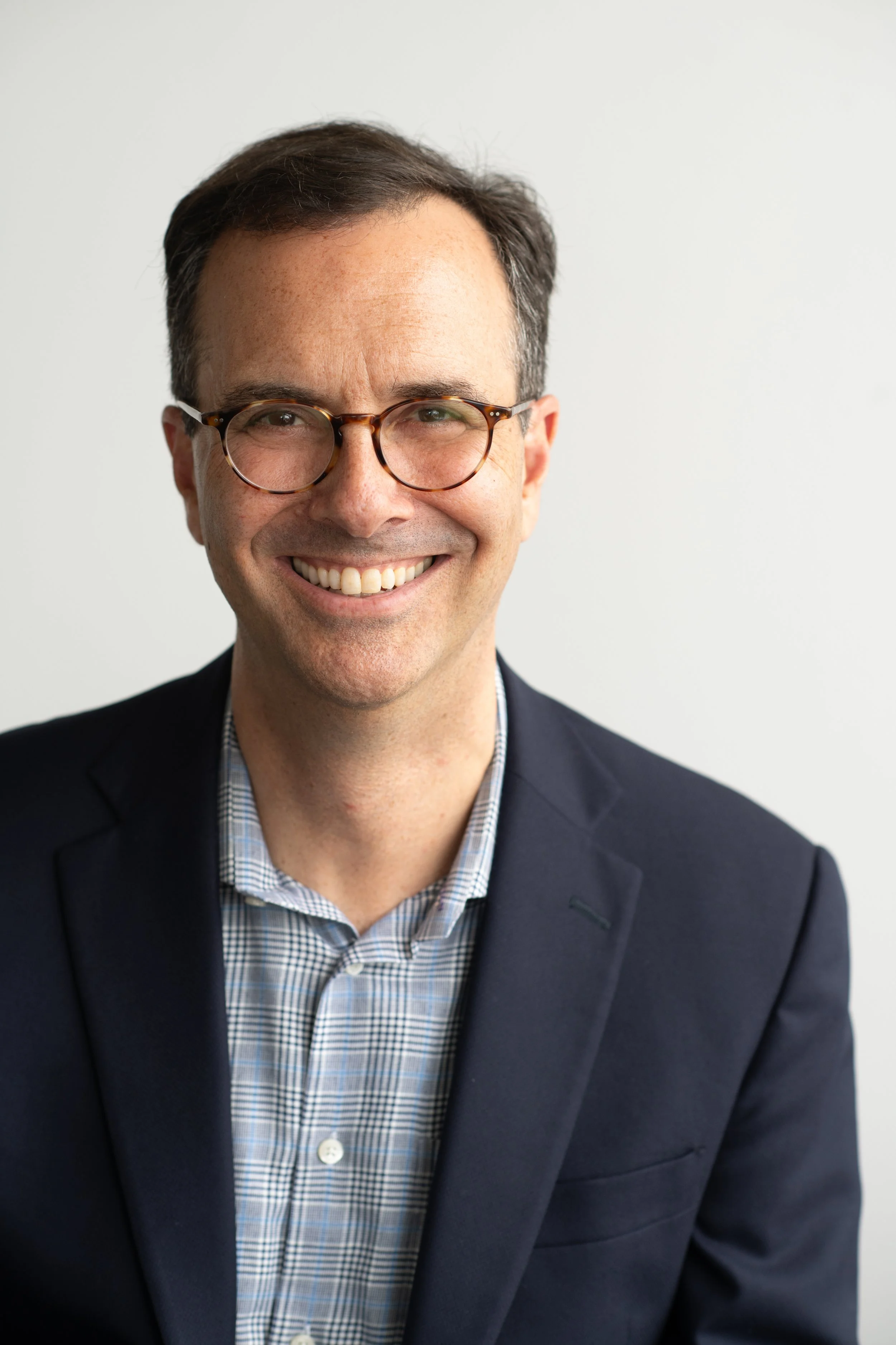 Headshot of a smiling man wearing glasses, a checked shirt, and a dark blazer against a plain white background.