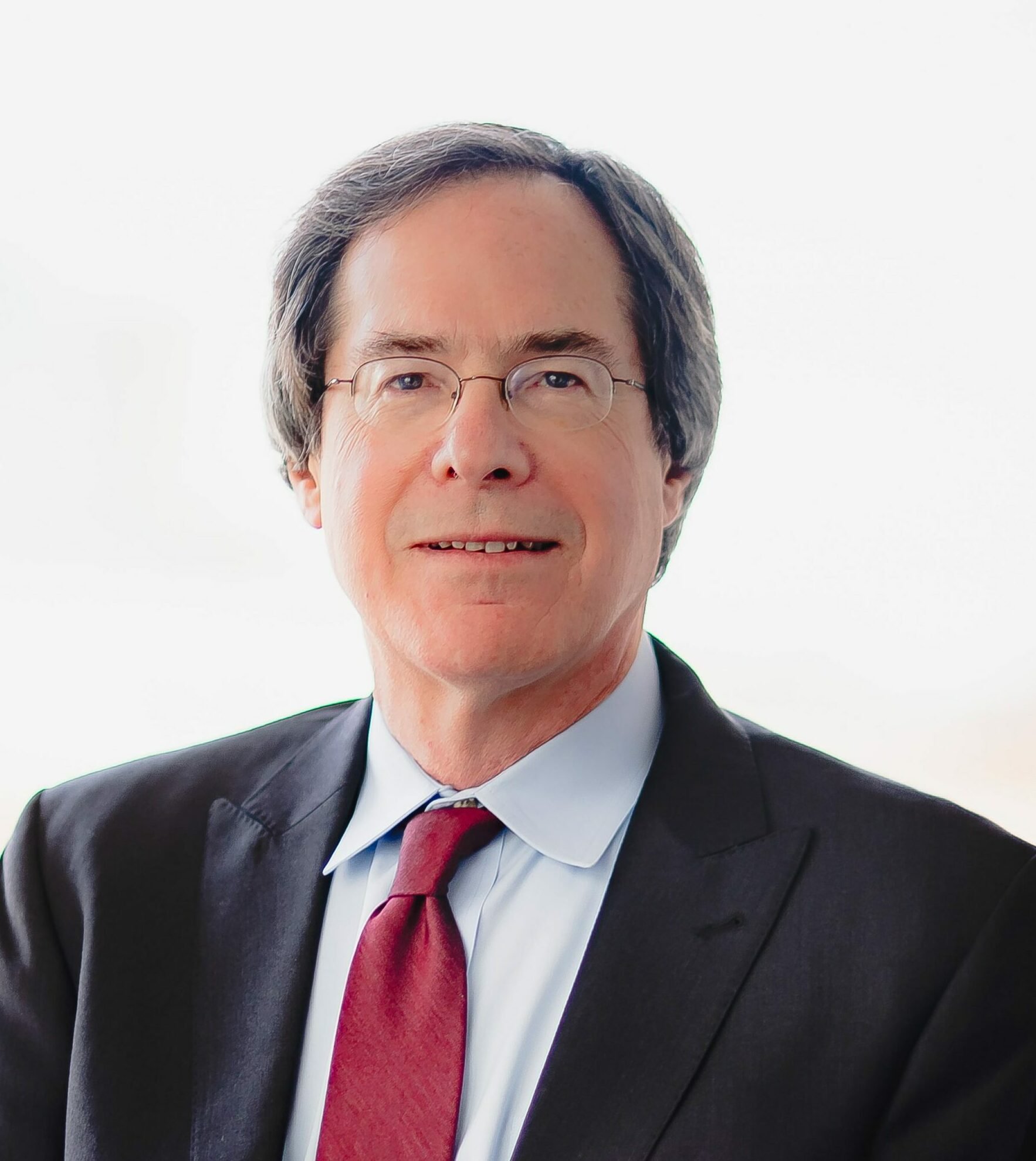 Close-up of a man wearing glasses, black suit, white shirt, and red tie, with a blurred white background.