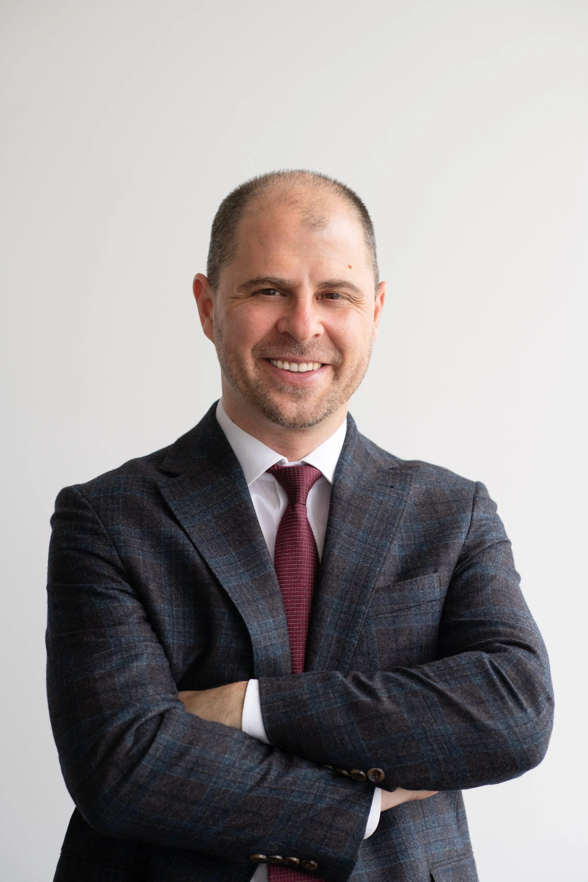 A man with a close-shaved head and beard, smiling with arms crossed, wearing a dark plaid suit and maroon tie, against a plain white background.
