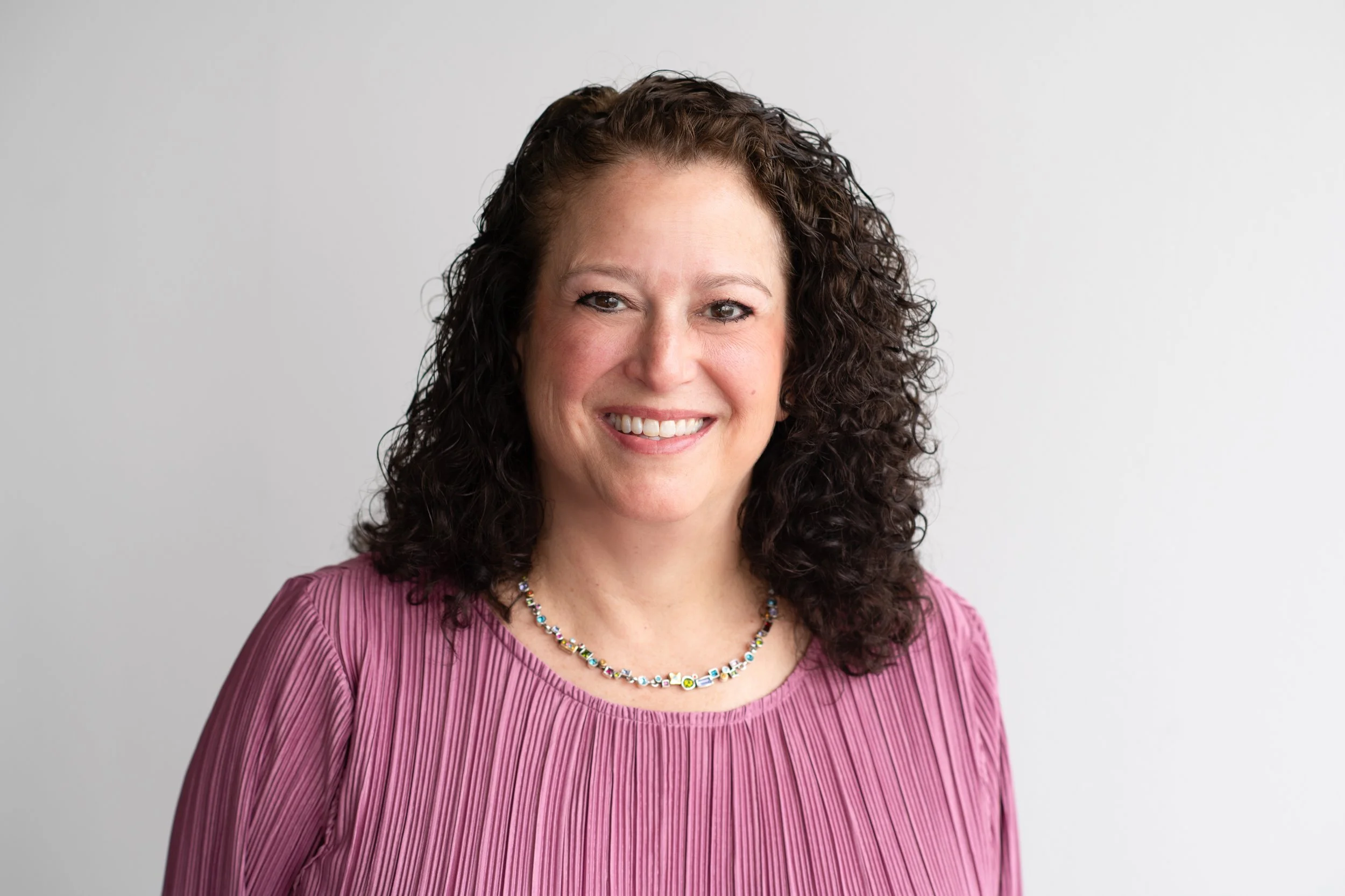 A smiling woman with curly brown hair wearing a pink blouse and multicolored beaded necklace against a plain white background.