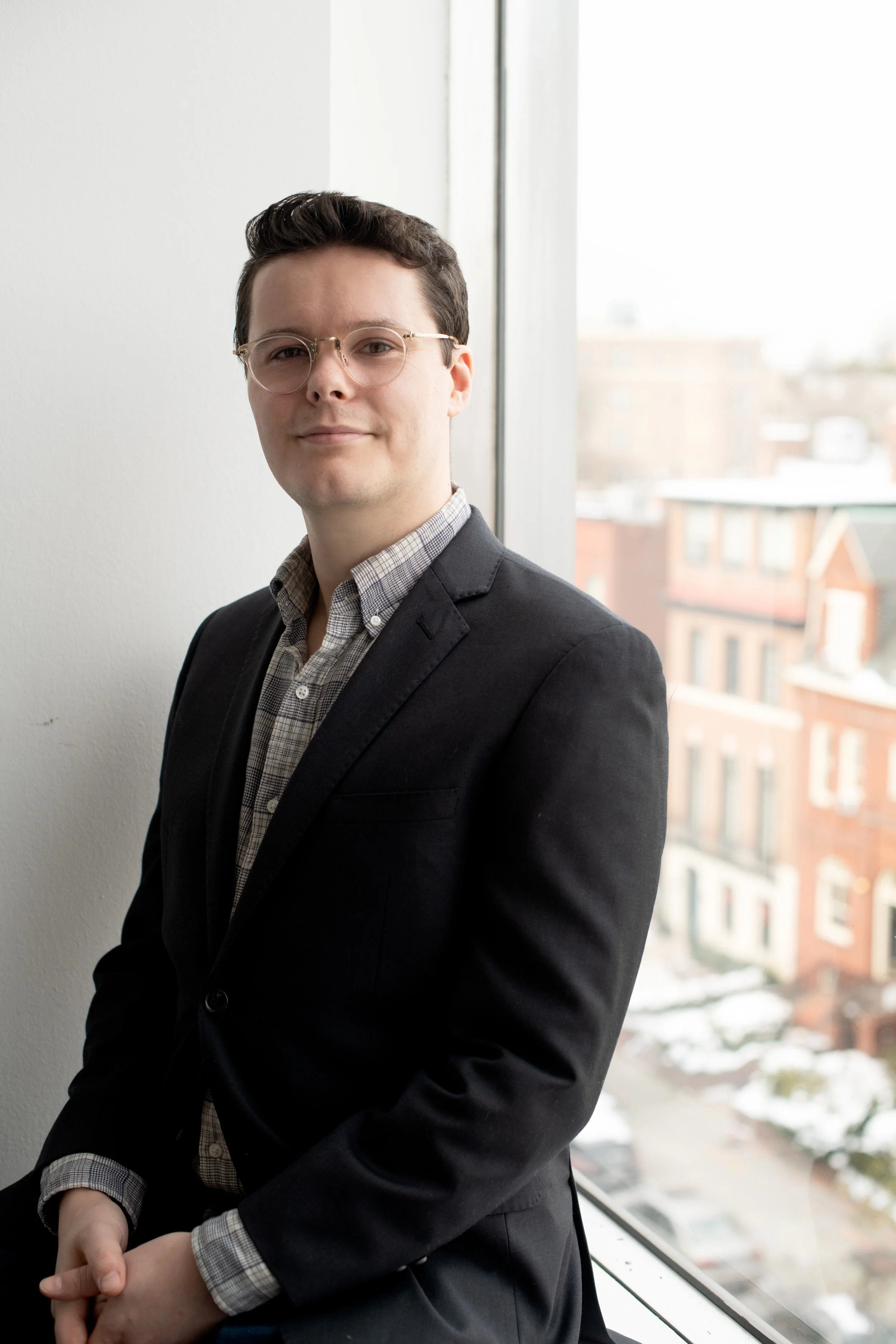 A young man with short dark hair and glasses, dressed in a dark blazer and a checkered shirt, standing indoors near a window with a cityscape in the background.