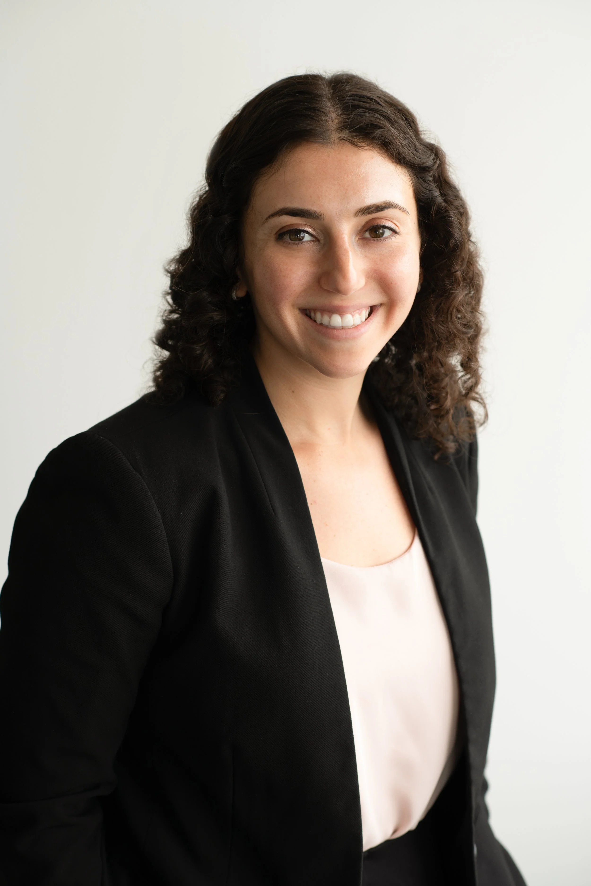 A woman with dark curly hair wearing a black blazer and light pink top, smiling against a white background.
