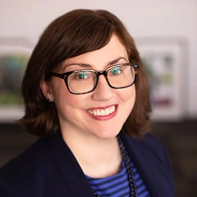A woman with brown hair and glasses smiling, wearing a dark blazer and a blue striped top, in an indoor setting.