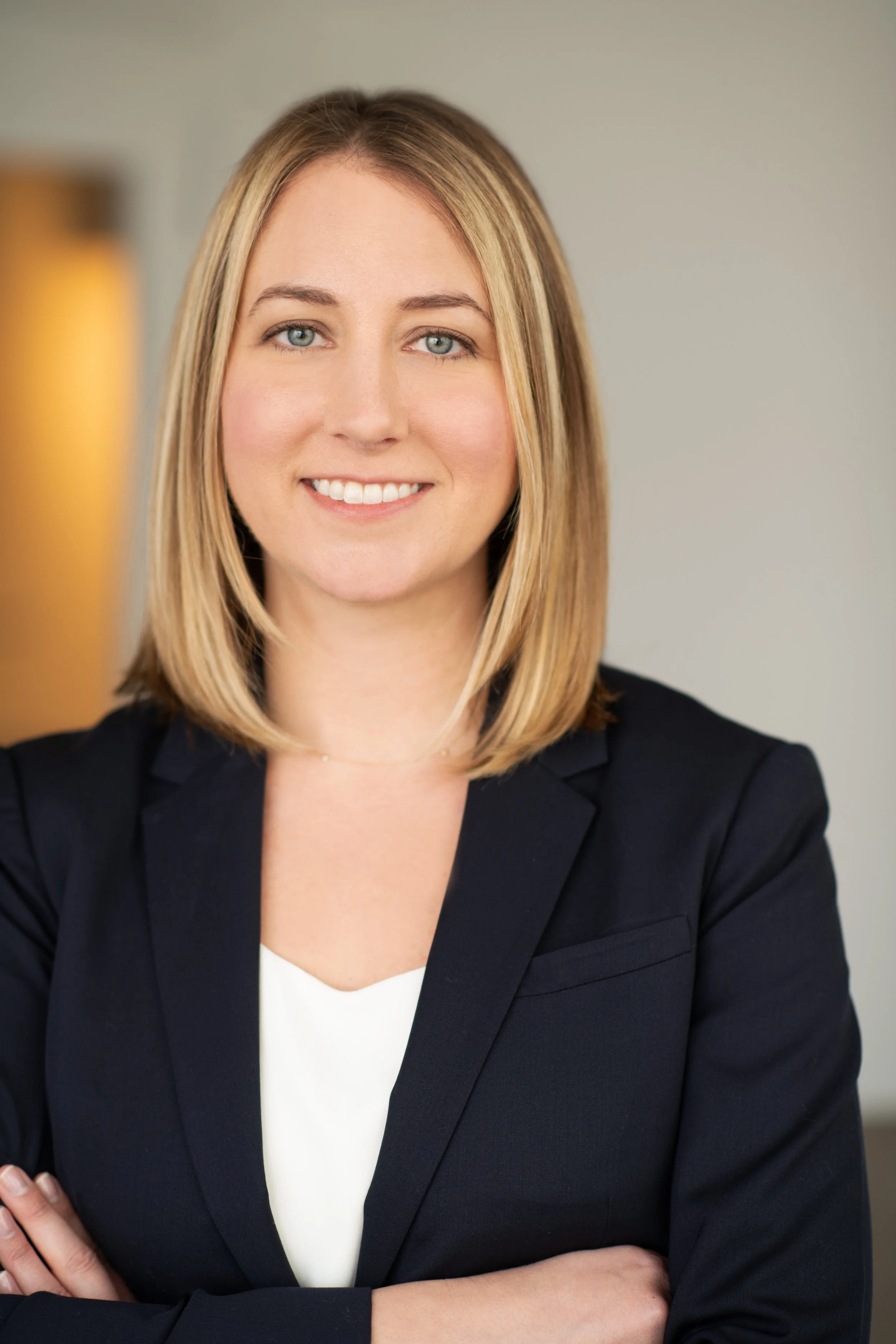 Close-up of a woman with blonde hair and blue eyes, smiling, wearing a black blazer and white top, posing with crossed arms.