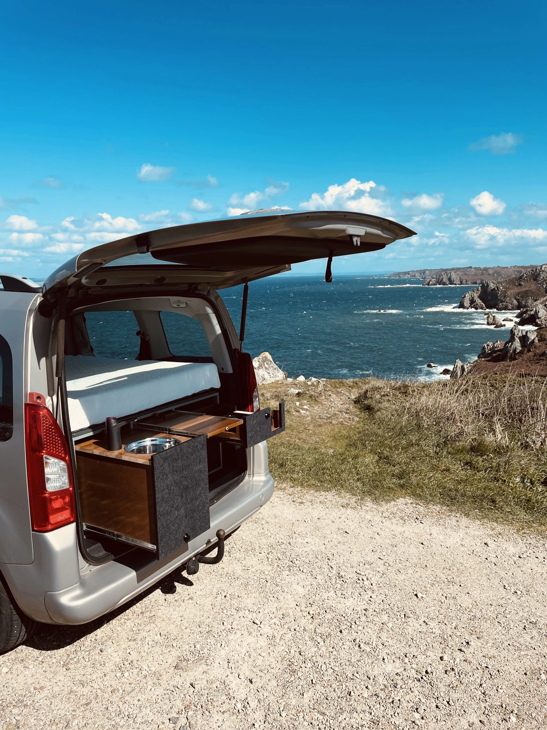 Un véhicule utilitaire blanc avec un toit ouvert, aménagé comme cuisine de camping, garé près de la côte avec vue sur la mer, des rochers et un ciel bleu avec quelques nuages.