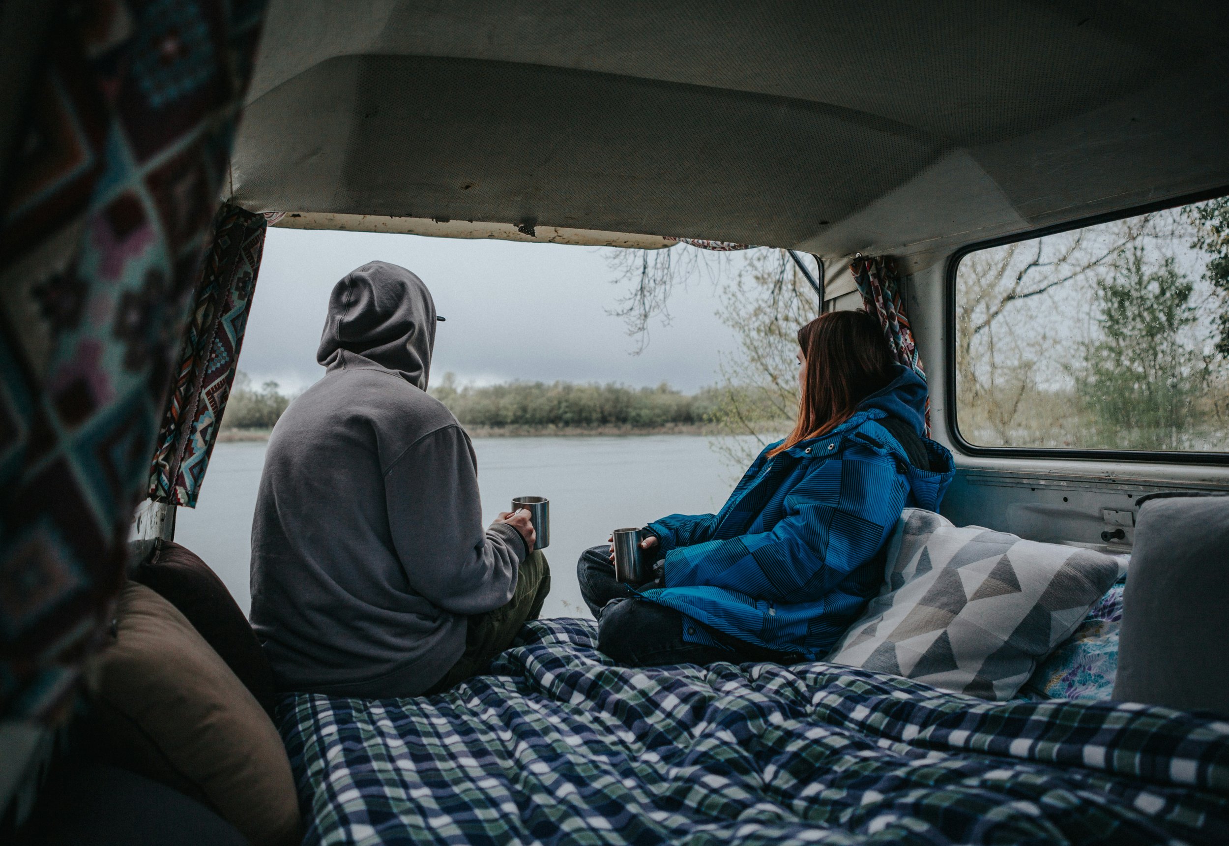 Deux personnes assises dans un van en regardant dehors, une personne en hoodie gris et une autre en veste bleue, avec vue sur un lac ou une rivière et des arbres.