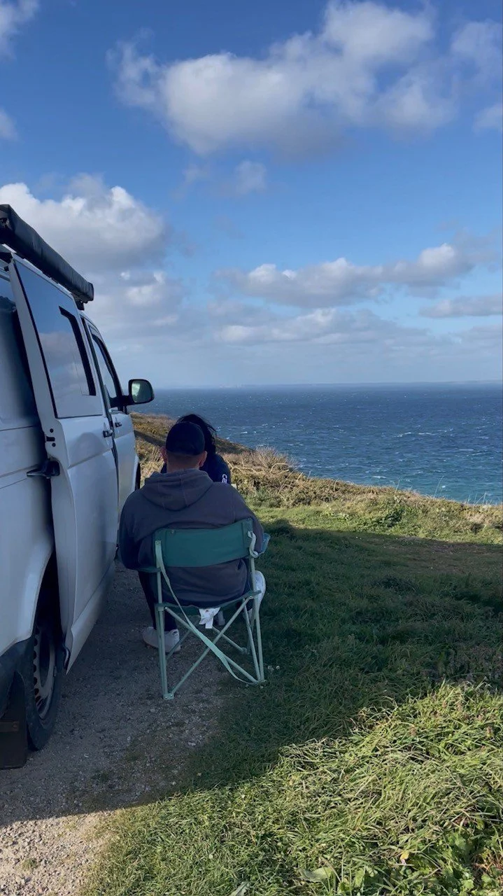 Personne assise sur une chaise pliante près de la côte, regardant l'océan, avec un véhicule blanc à côté, ciel partiellement nuageux.