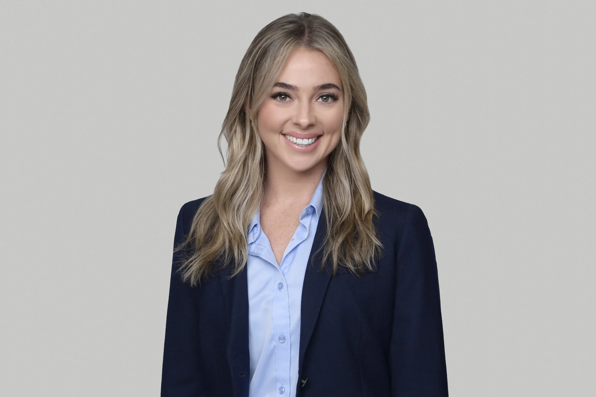 A woman with long blonde hair wearing a navy blazer and light blue shirt, smiling against a plain gray background.