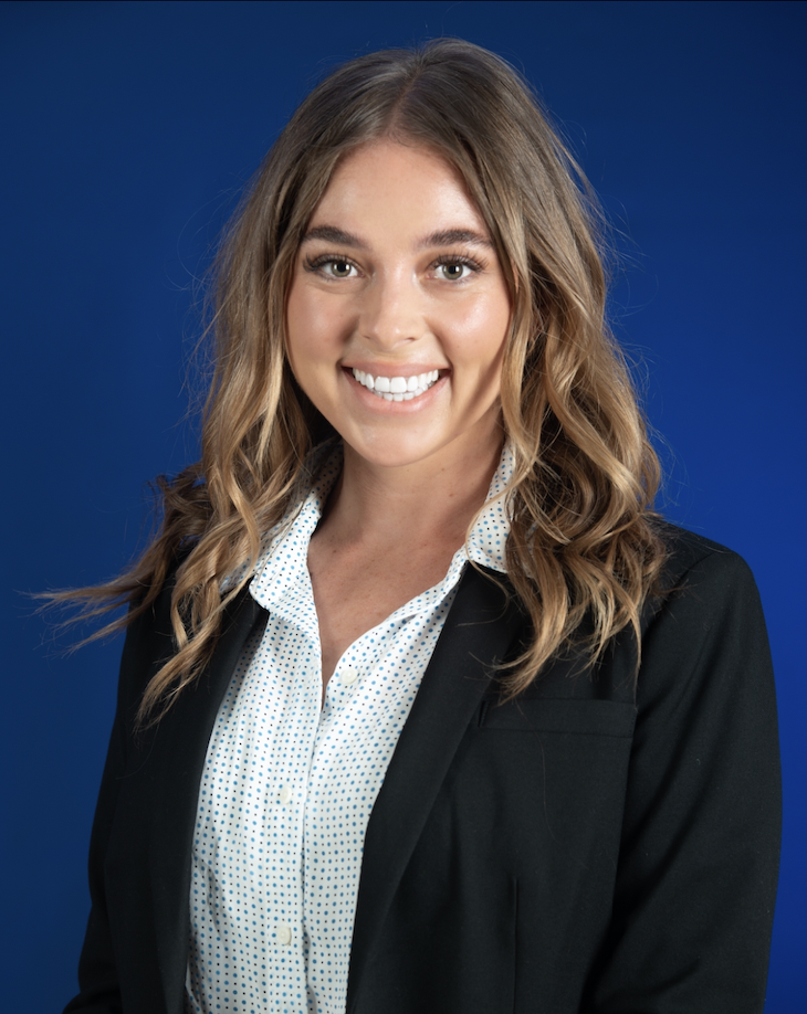 Professional woman with wavy blonde hair, wearing a black blazer and white blouse with small blue polka dots, smiling against a blue background.