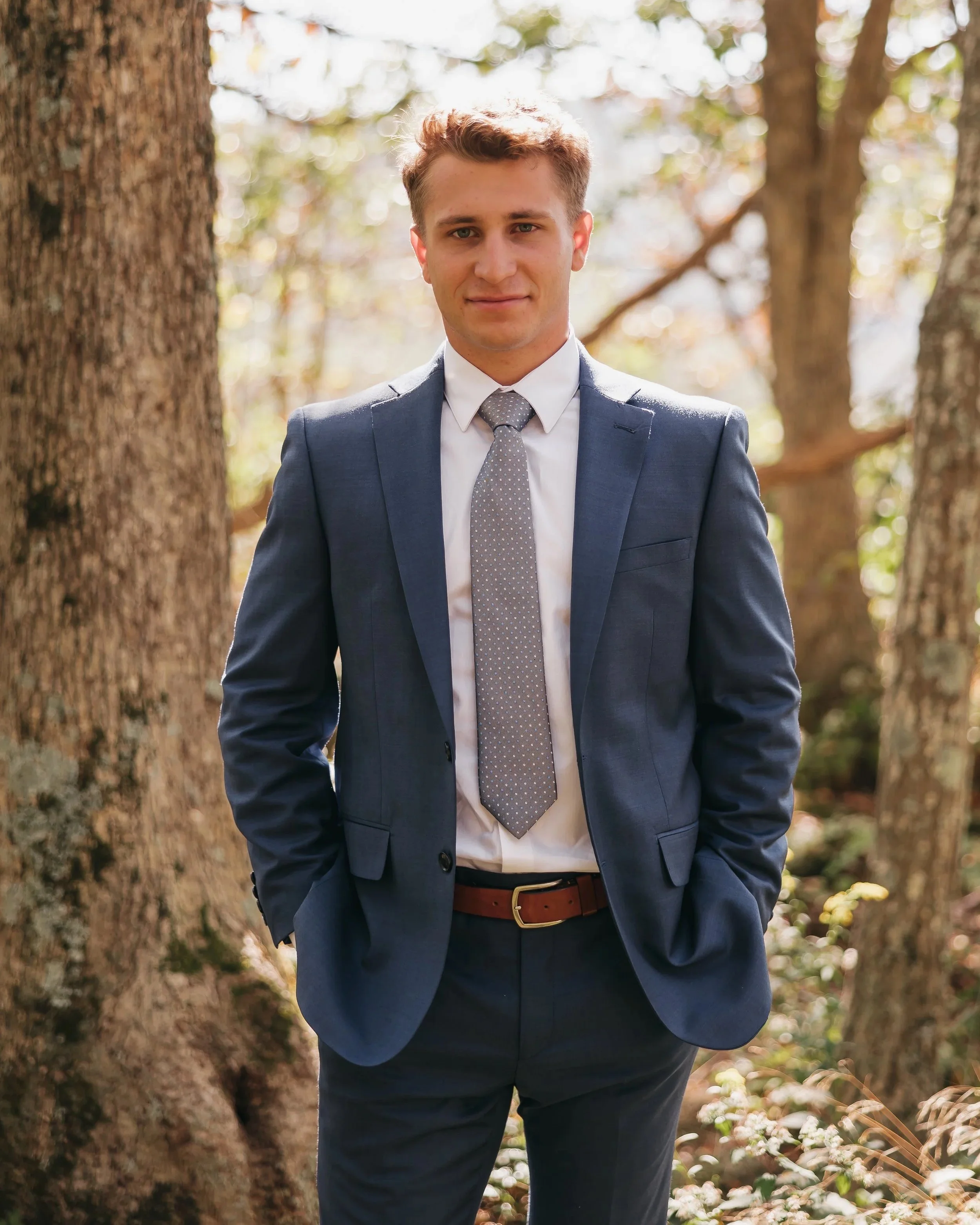 A young man with light brown hair and blue eyes wearing a dark gray suit, white shirt, and gray tie, standing outdoors between large trees with green and brown leaves. He is smiling slightly and looking at the camera, with his hands in his pockets.