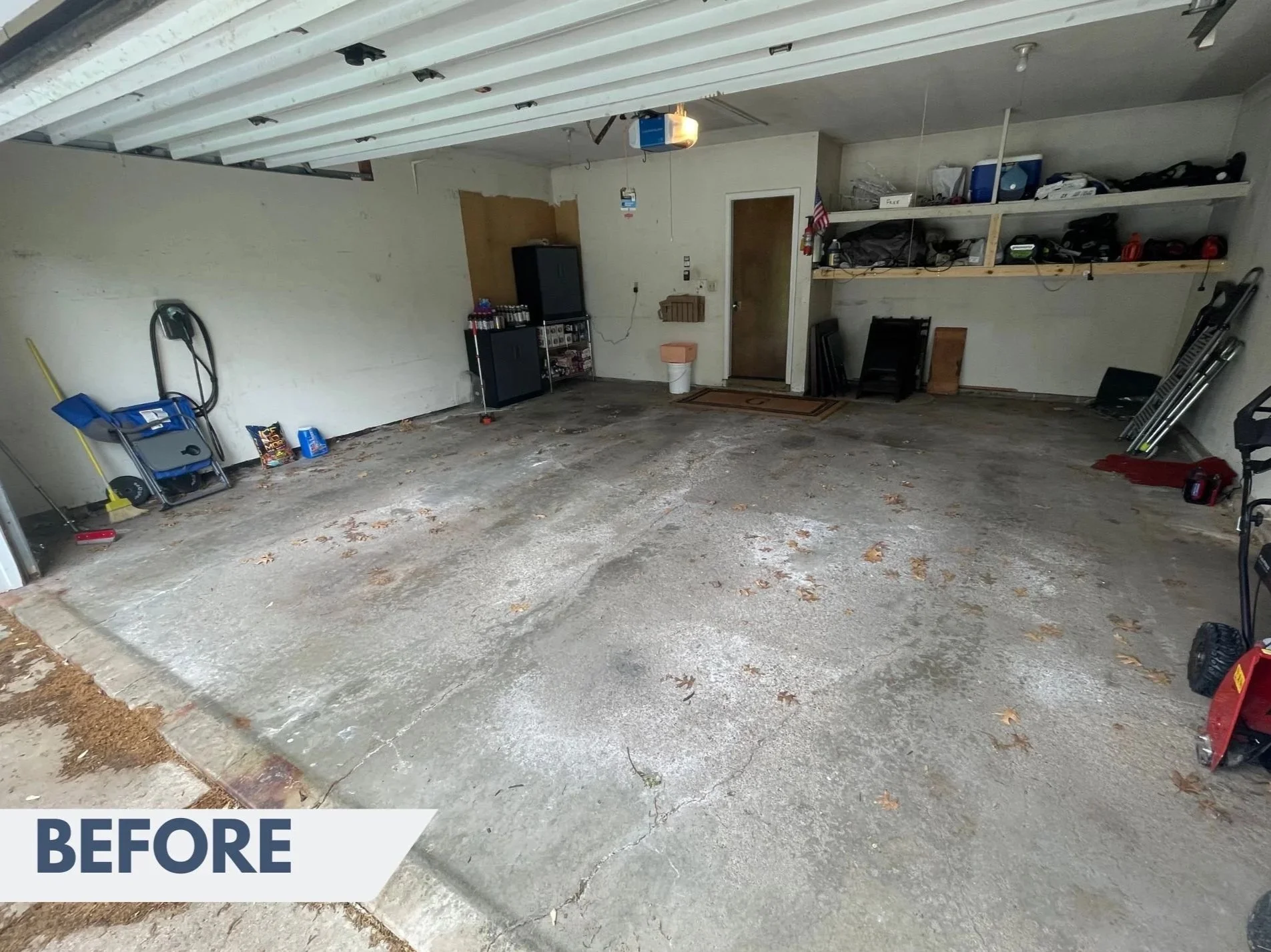 An empty garage with a concrete floor and a ceiling with garage door tracks, items stored on shelves, and some tools and supplies on the ground.