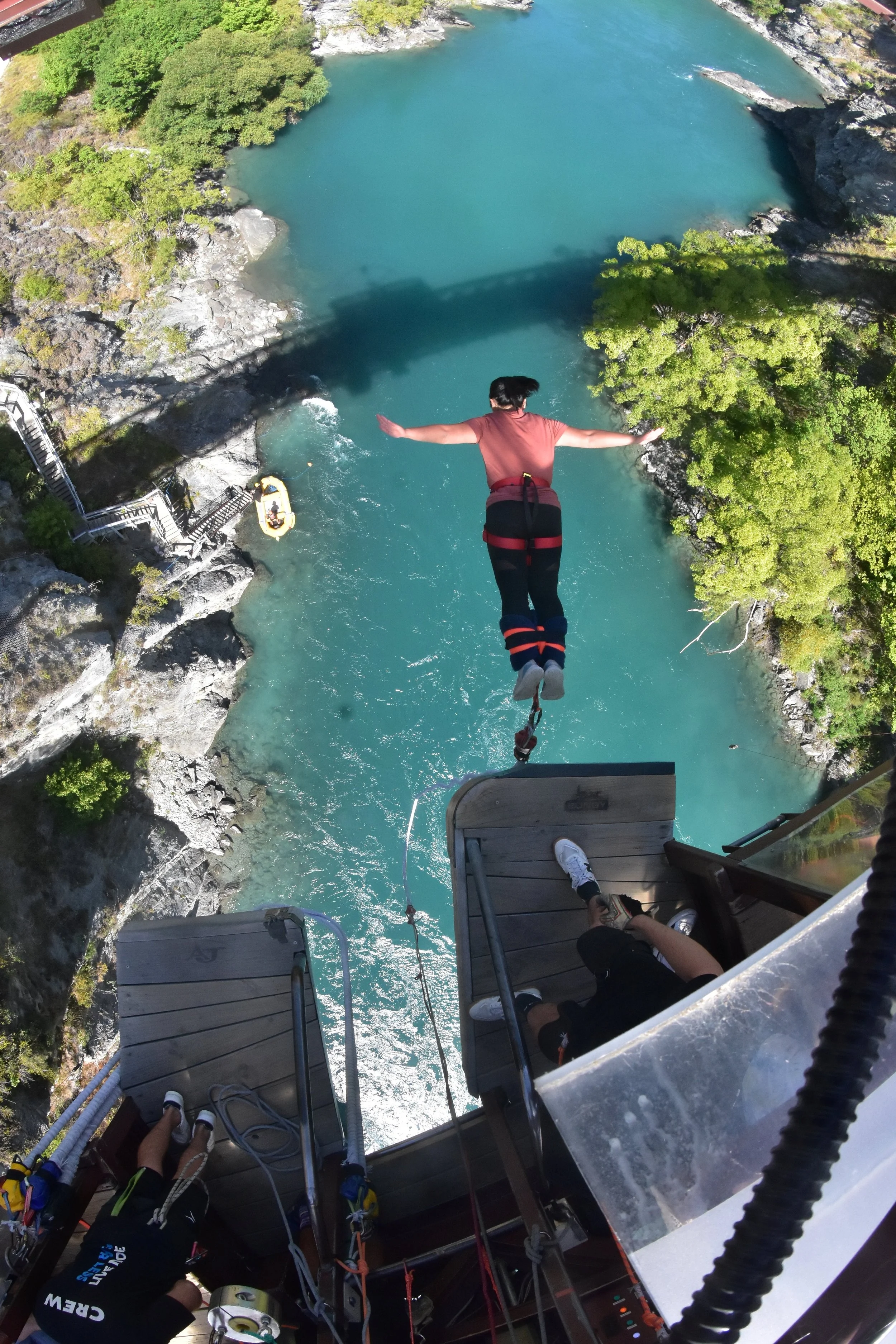 A person bungee jumping from a platform over a river with rocky banks and green trees, with other people visible sitting on the platform.