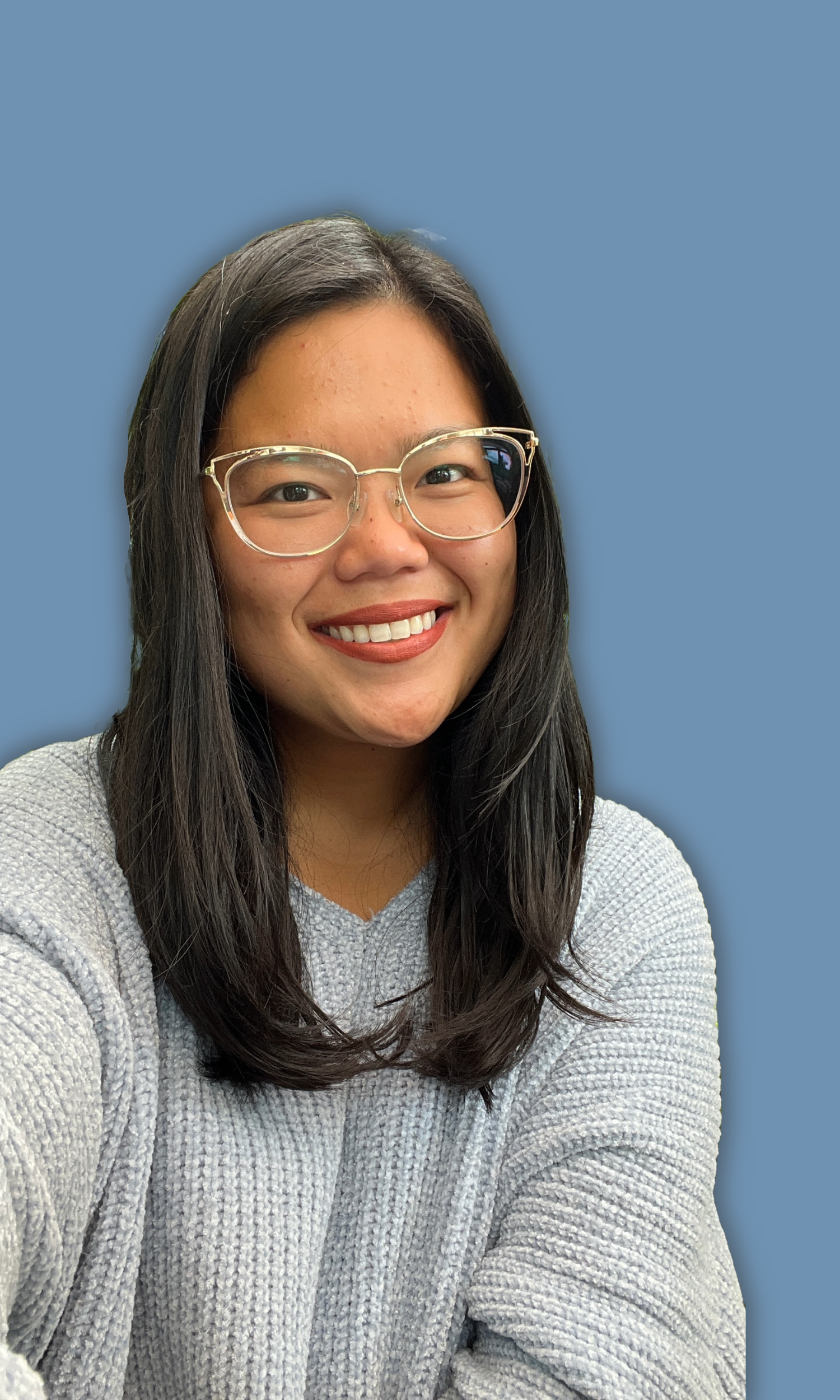 A smiling young woman with long dark hair, wearing clear glasses and a light gray sweater, sitting against a plain blue background.