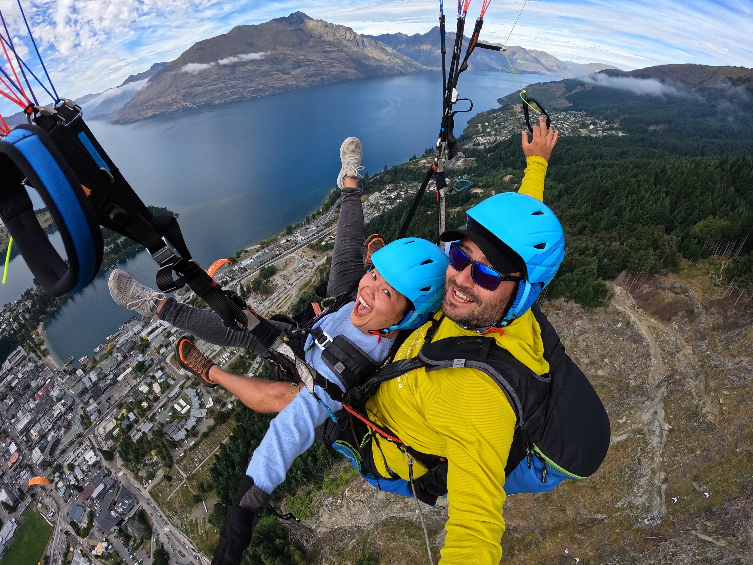 Two people paragliding over a city and a lake with mountains in the background. One person is wearing a blue helmet and sunglasses, and the other person is wearing a yellow jacket and a blue helmet. The city below has a grid pattern with roads and buildings.