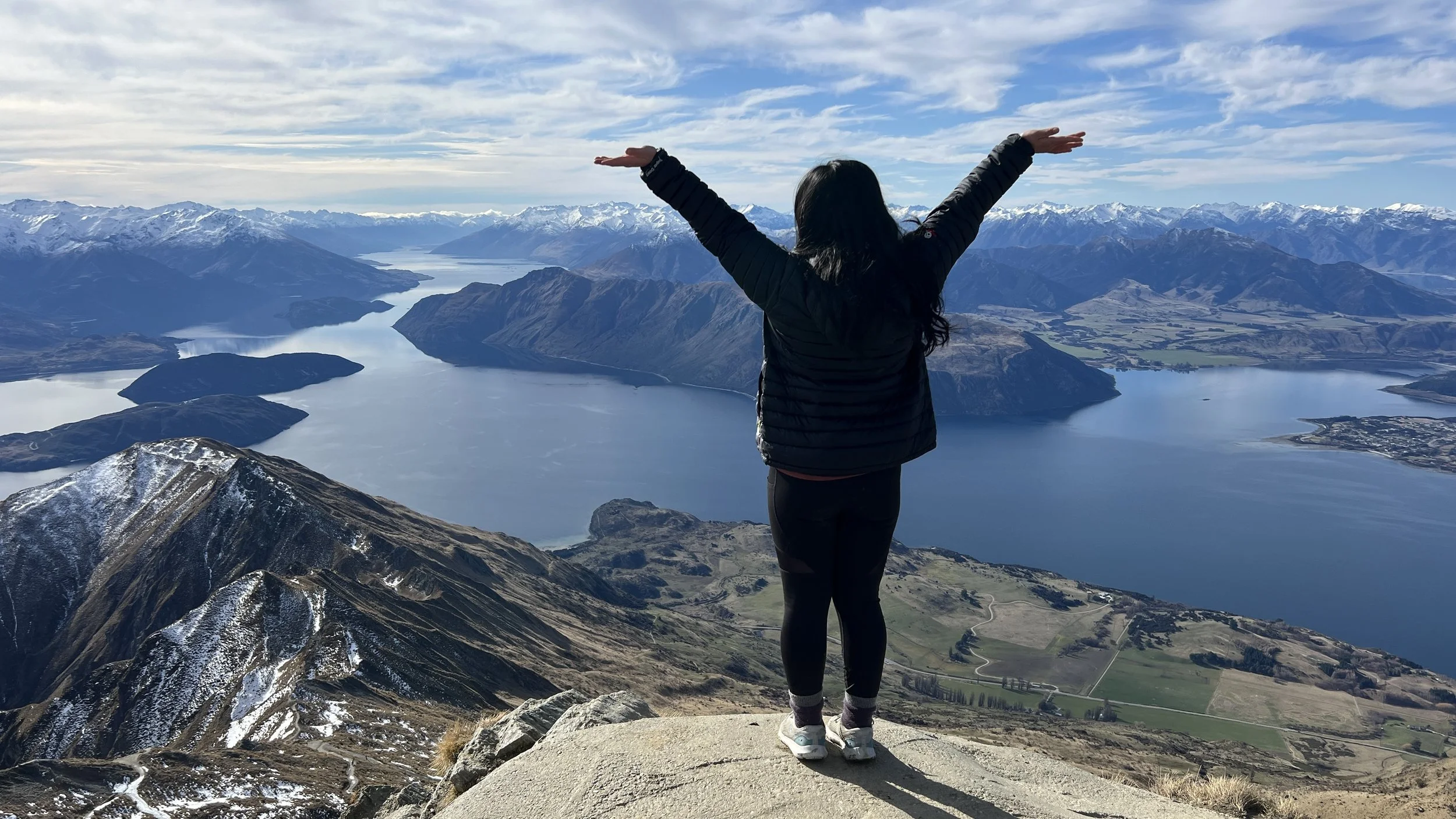 A person standing on a mountain peak with arms outstretched, overlooking a lake surrounded by snow-capped mountains under a partly cloudy sky.