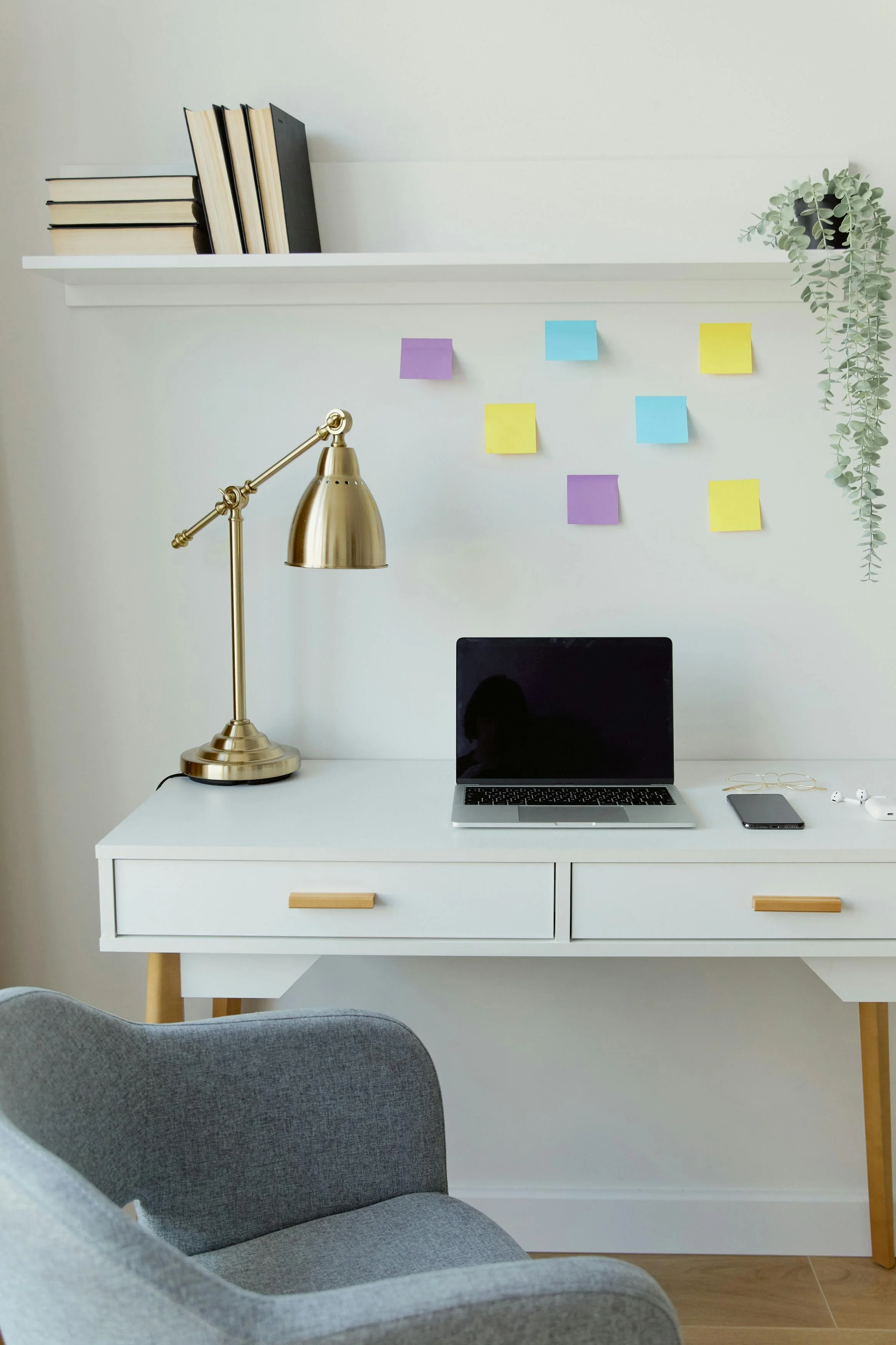 A white desk with a gold desk lamp, a laptop, smartphone, and glasses. Above the desk, a white shelf with books and sticky notes, and a potted plant hanging down on the right. In the foreground, a gray upholstered chair. The environment appears to be a home office or workspace.