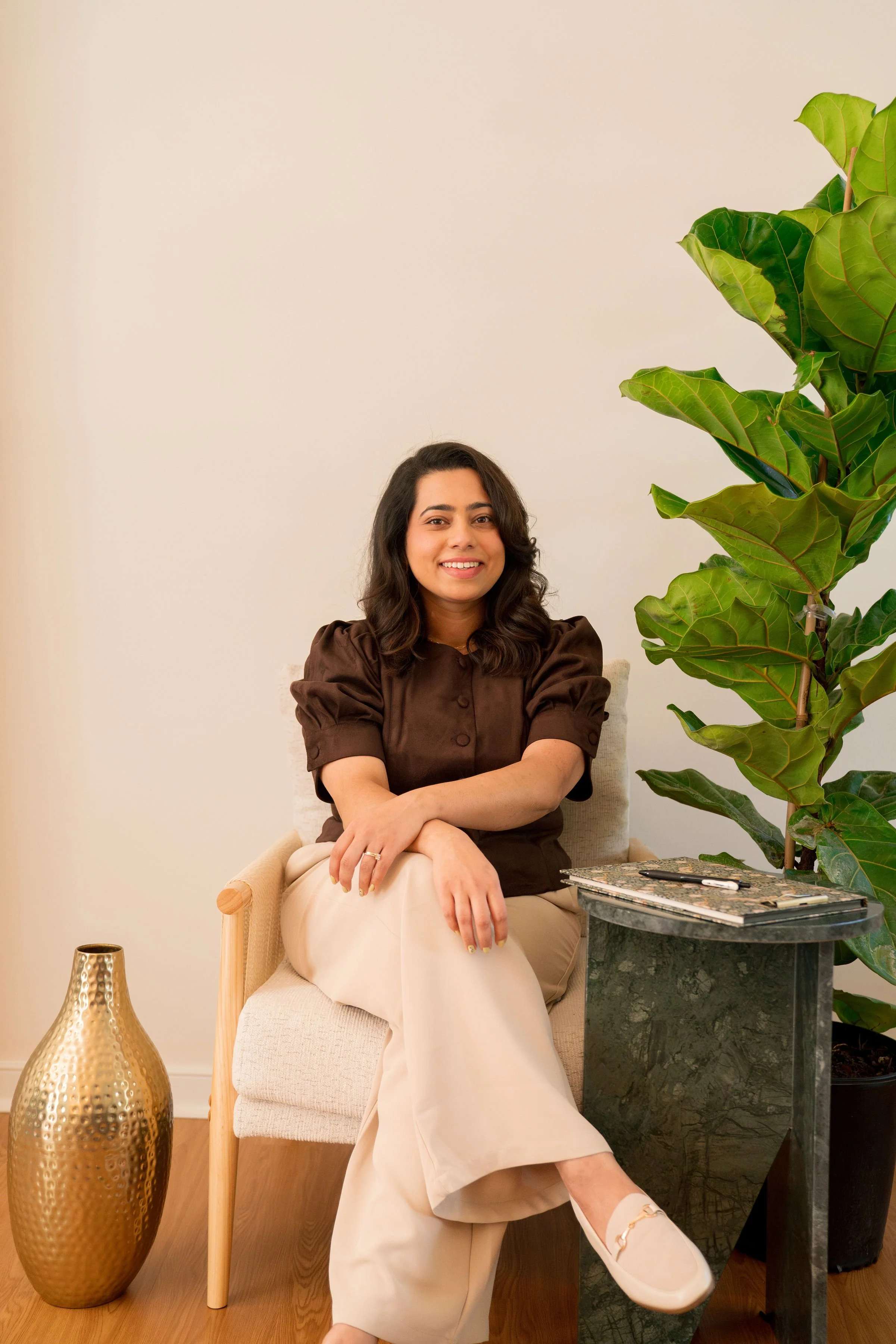 A woman with dark wavy hair smiling, sitting on a beige armchair. She is wearing a brown top and cream-colored pants, with her arms crossed. There is a tall green plant next to her, a black marble side table with a notebook and pen, a gold textured vase on the floor, and a gold decorative object on the table on her left, with a plain light-colored wall background.