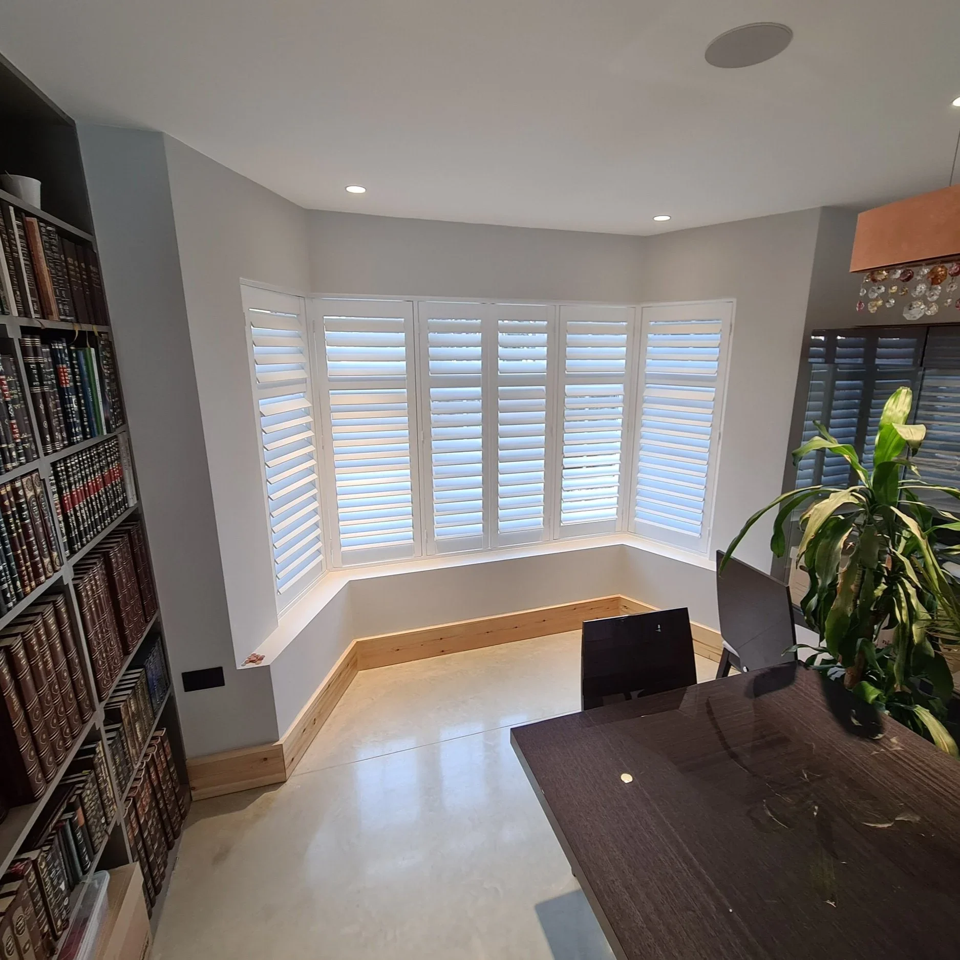 A room with large bay windows featuring white plantation shutters, a piece of dark wood furniture, a tall bookshelf filled with books, a potted plant, and a hanging light fixture with decorative glass accents.