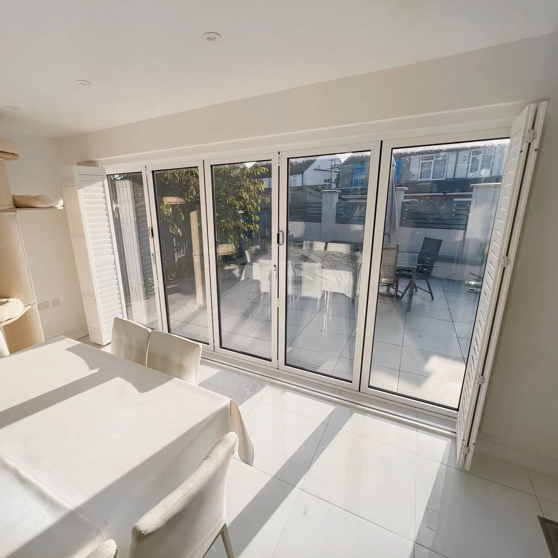 Living room with white walls, a white dining table with chairs, and large glass sliding doors leading to an outdoor patio with a table and chairs. Outside, there are neighboring houses and a tree.
