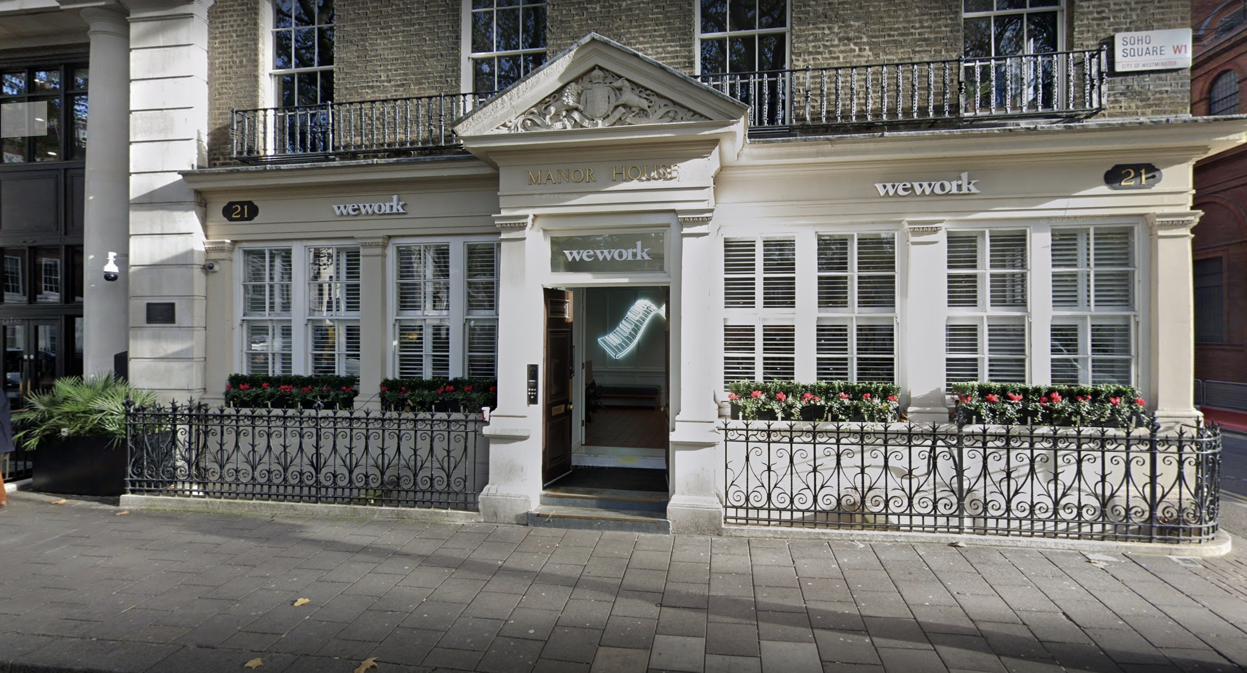 Front view of a building with a sign reading 'Manor House', adorned with flowers in window boxes and a neon ladder sign inside, with the street name 'Soho Square' visible in London.