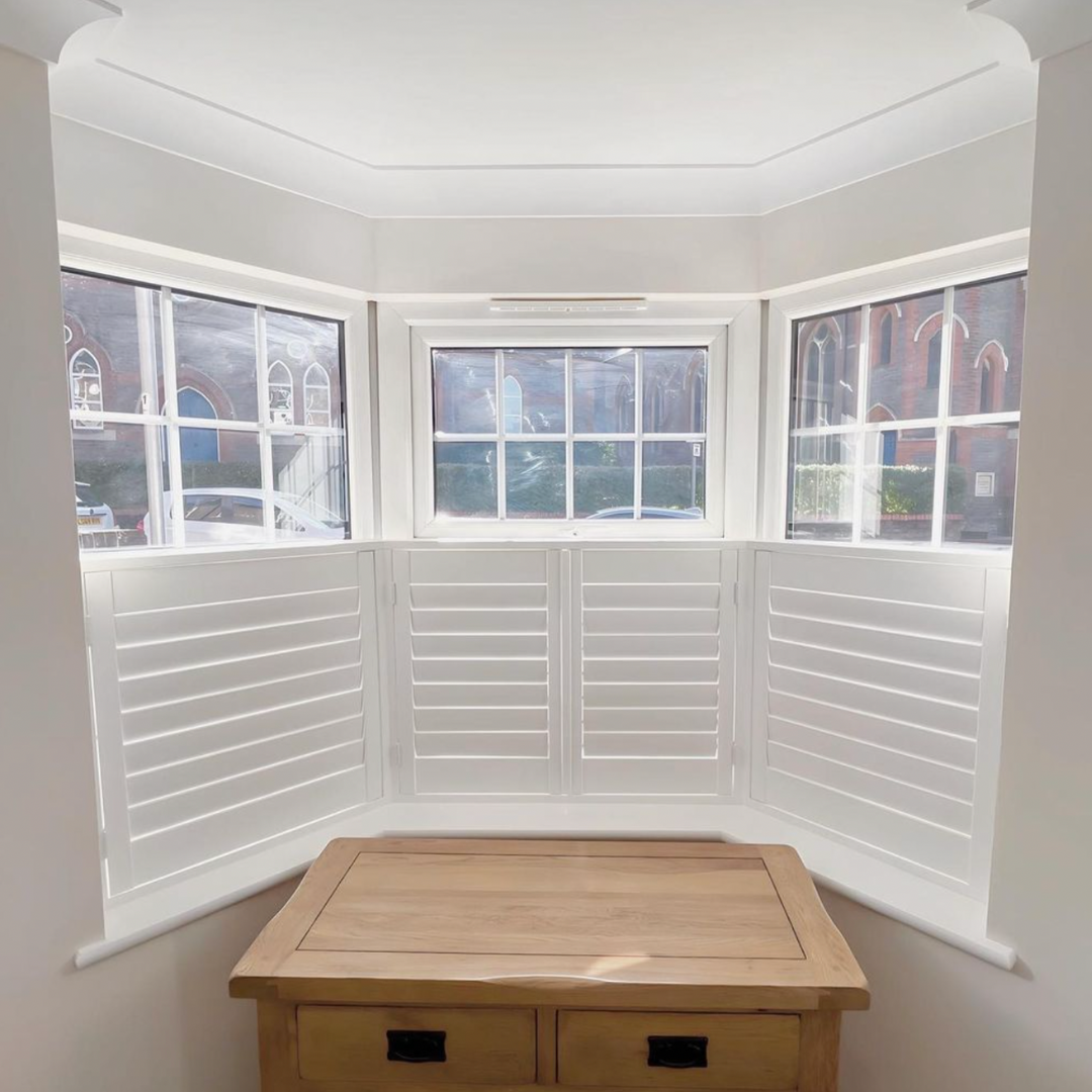 A bay window with white shutters and a wooden table underneath in a bright room.