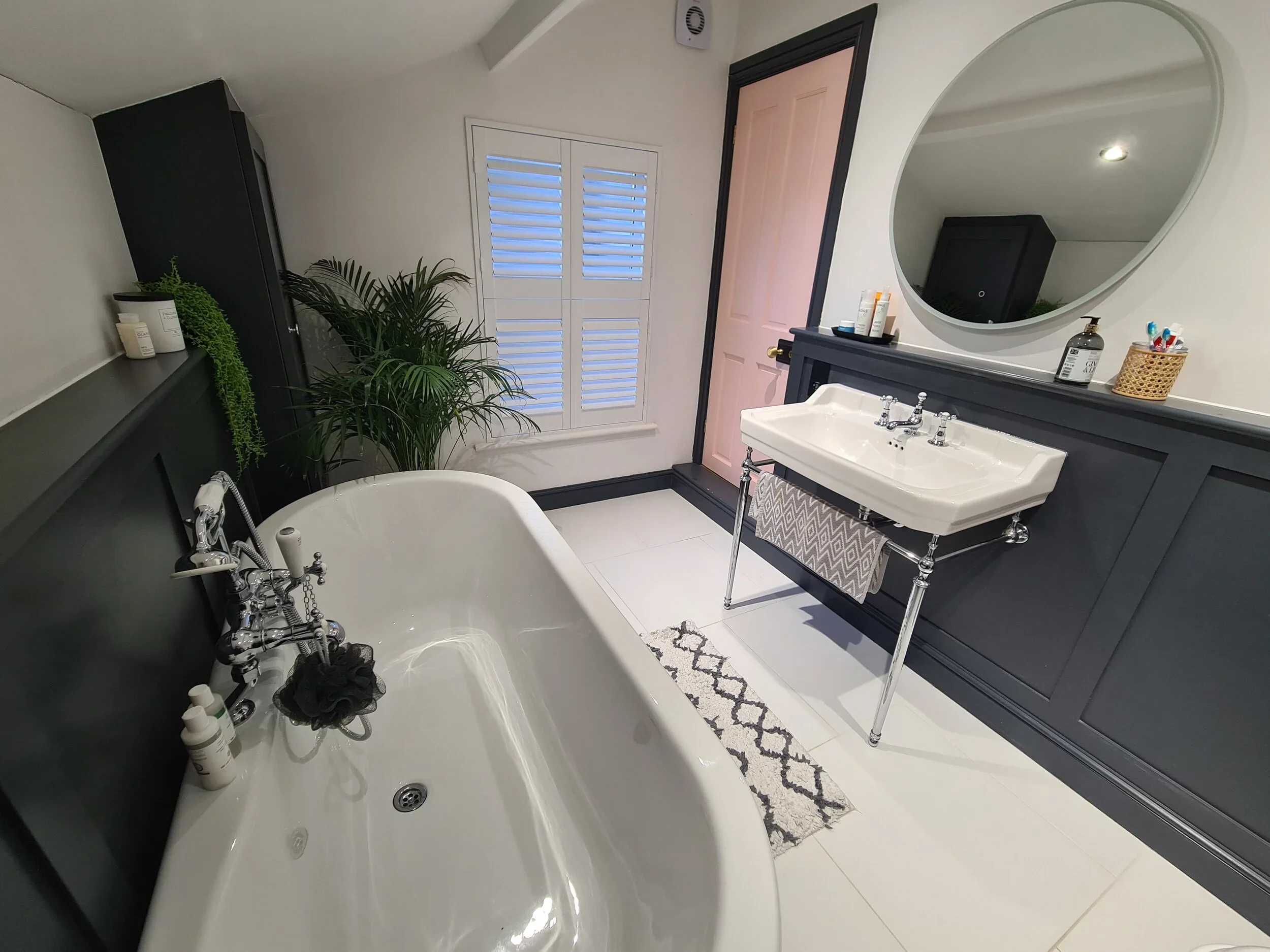 Bathroom with clawfoot bathtub, double sink vanity, large mirror, plant, window with shutters, closed door, and bathroom toiletries.