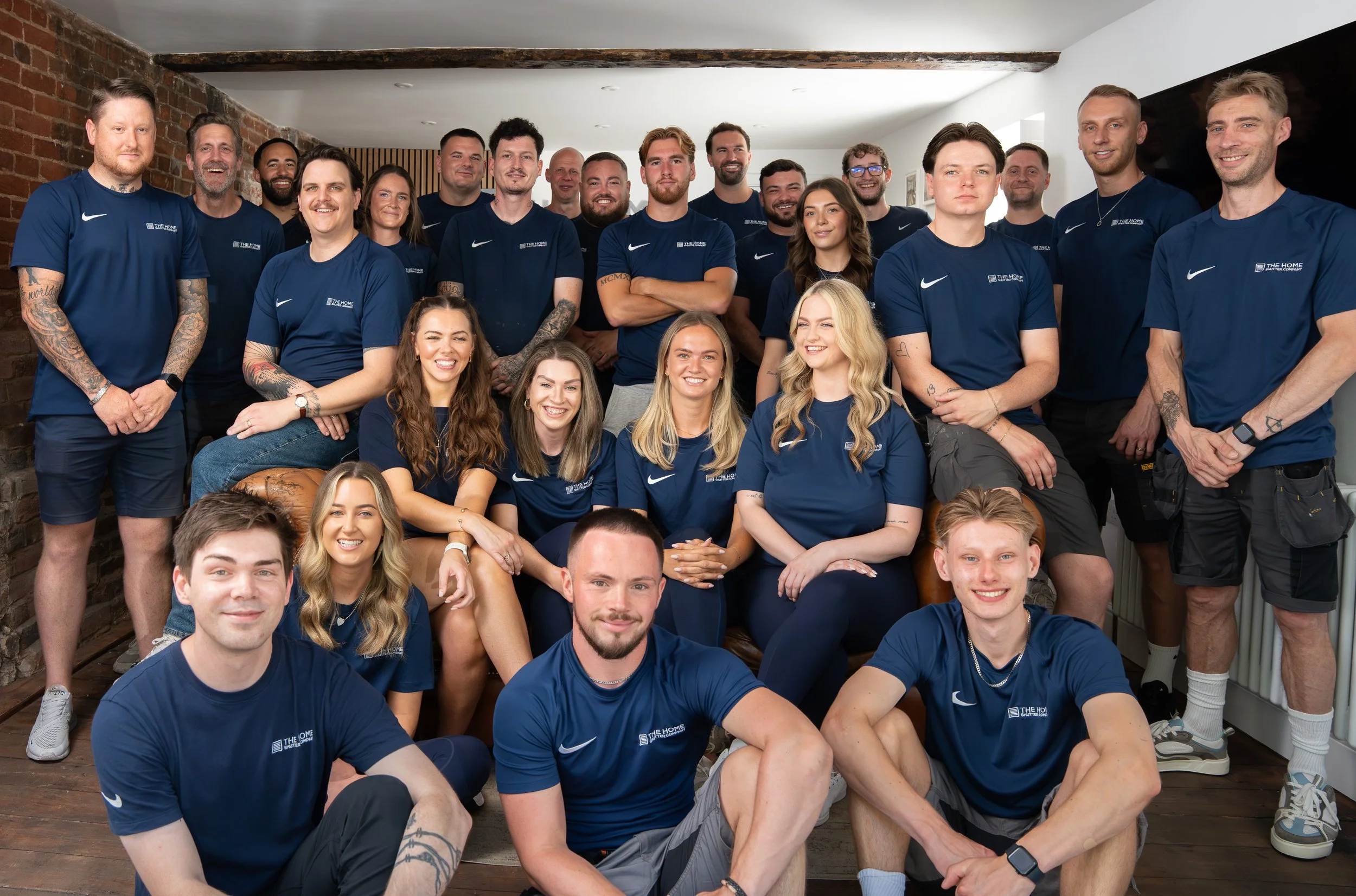 Group of people wearing matching blue shirts in an indoor space with brick and white walls, smiling for a group photo.