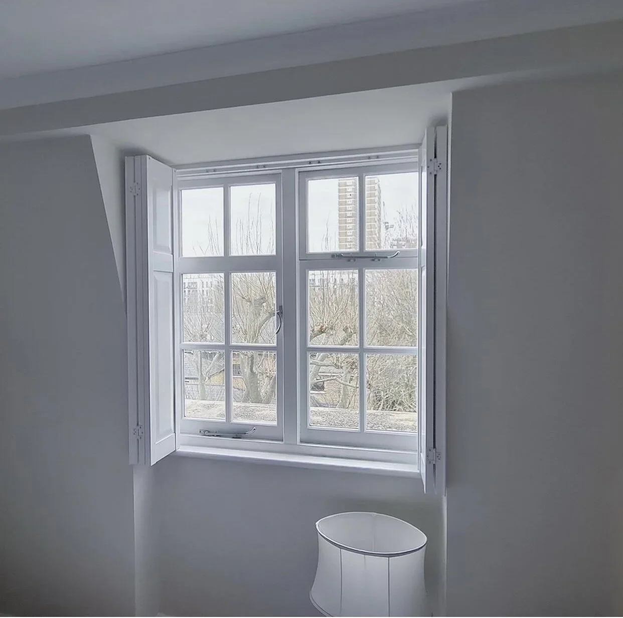 A window with white shutters, overlooking trees and a building in the distance, with a white lampshade below.