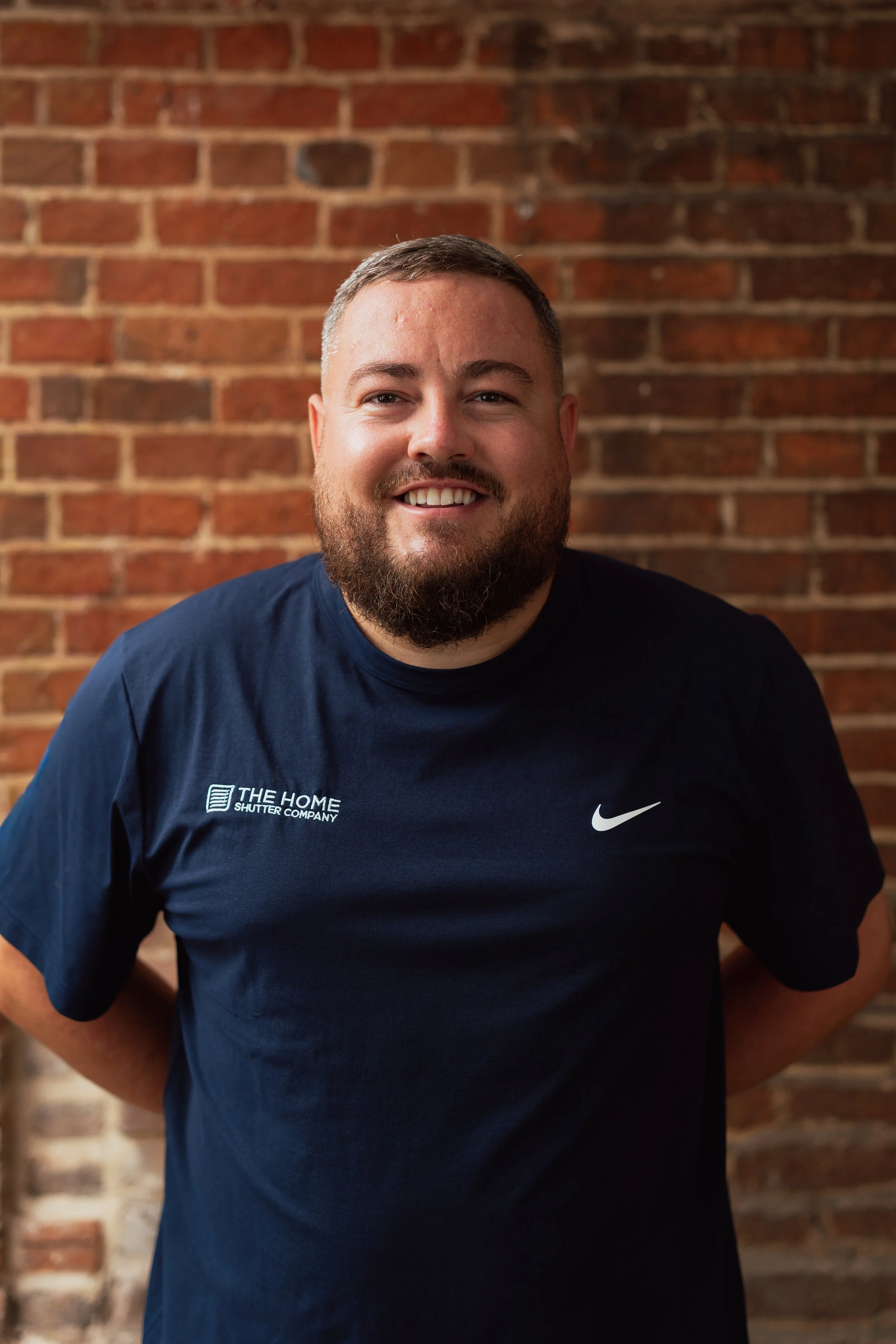 A smiling man with a beard and short hair wearing a navy blue Nike T-shirt standing in front of a brick wall.