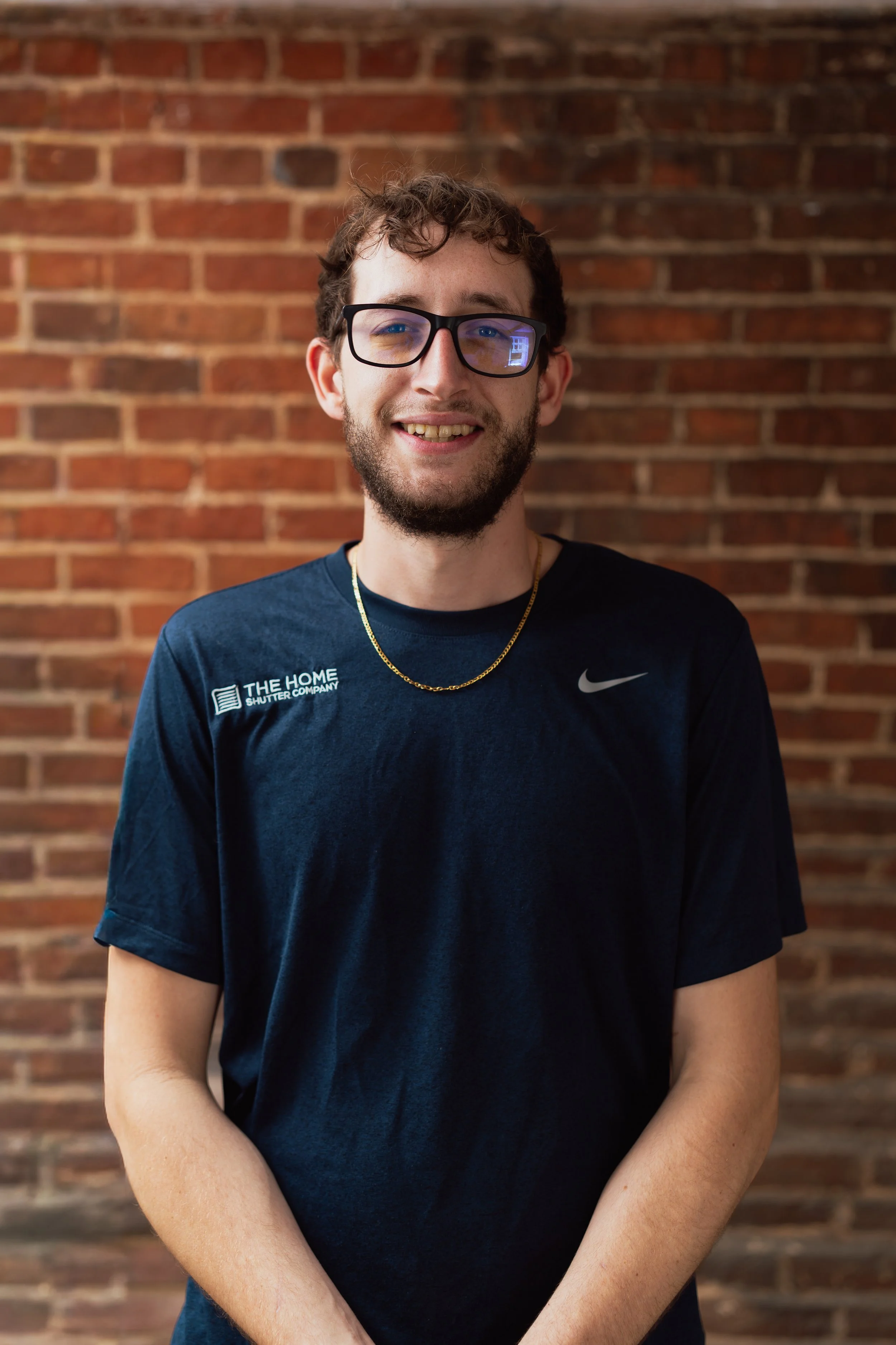 A young man with curly brown hair, glasses, and a beard, smiling, wearing a navy blue T-shirt and a gold chain, standing against a brick wall.