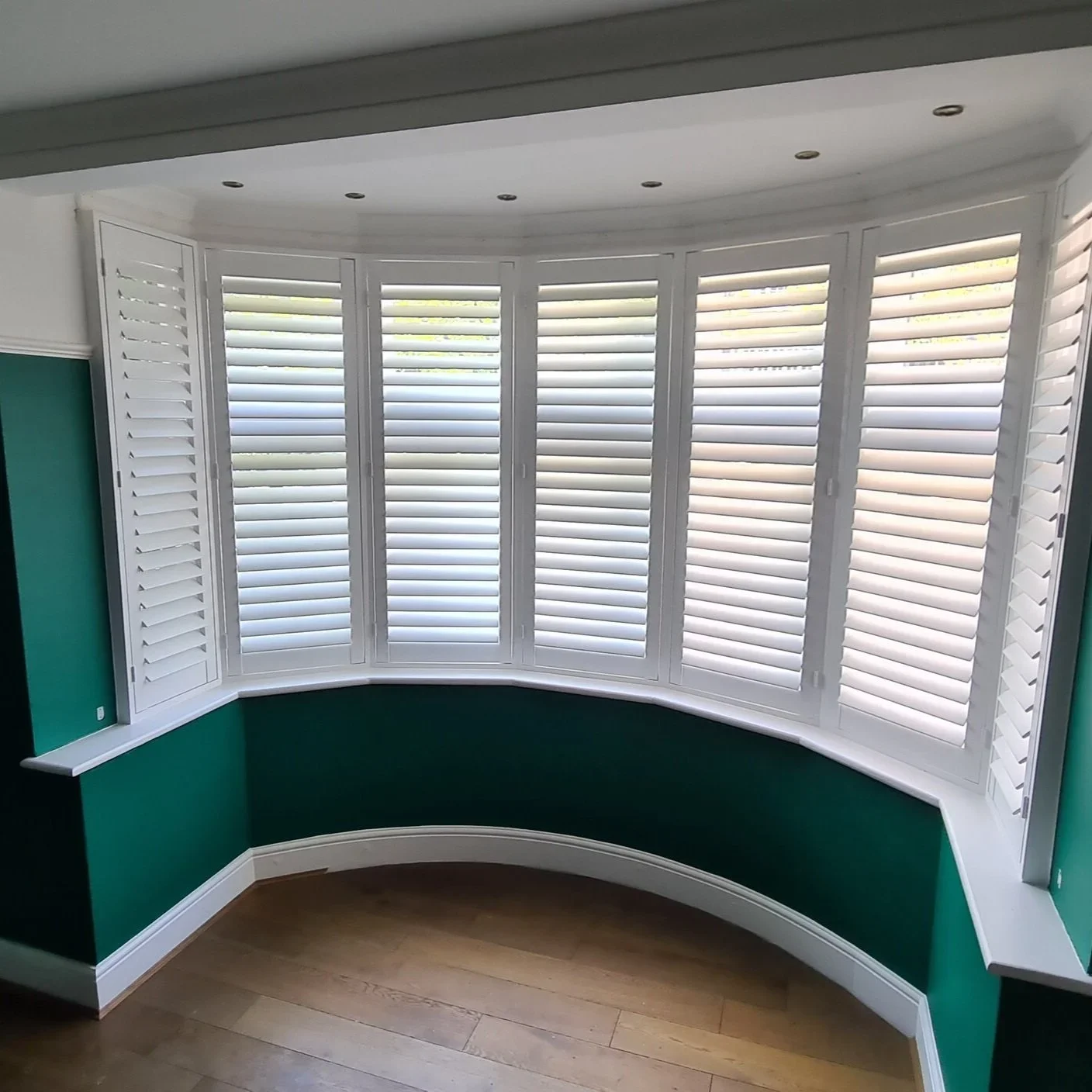 A curved wall with white plantation shutters covering a large window, green walls, hardwood flooring, and a white baseboard.