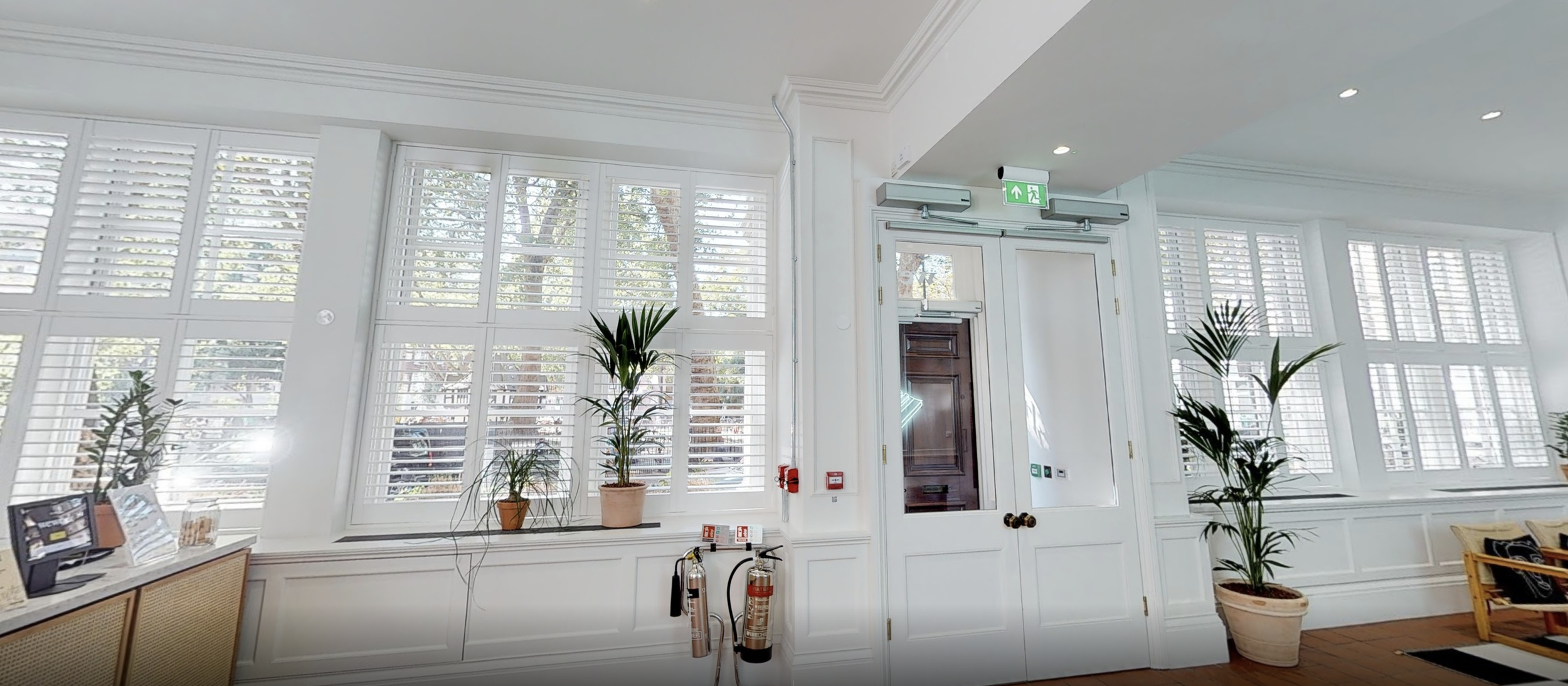 Interior of a bright lobby area with large windows with white shutters, potted plants, and a wooden door with an emergency exit sign above.