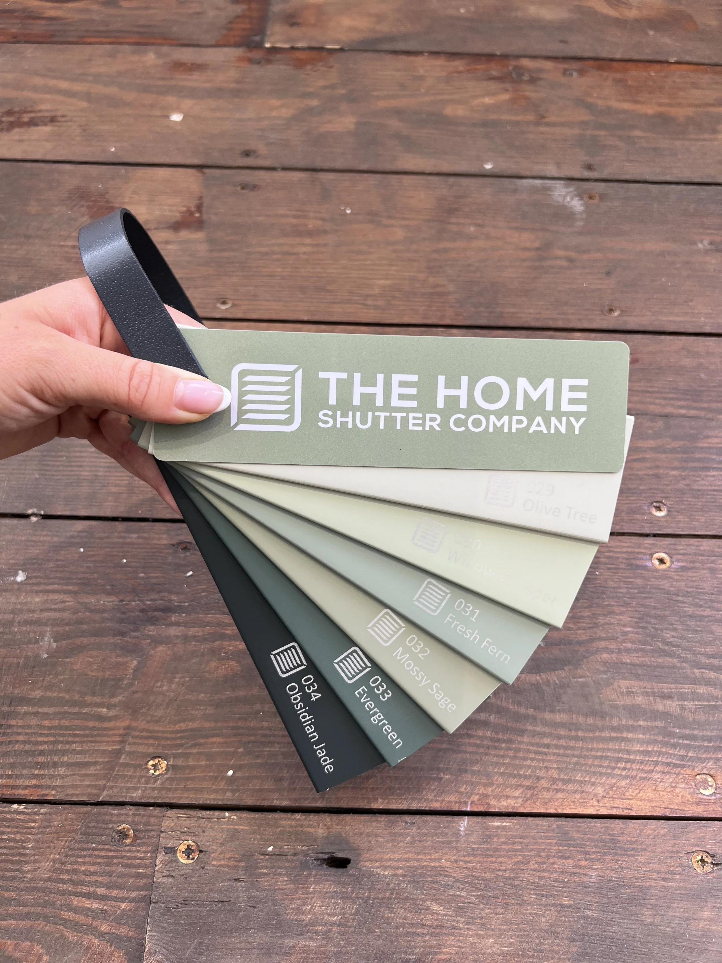 A hand holding a fan deck of color samples from 'The Home Shutter Company,' fanned out across a wooden surface.