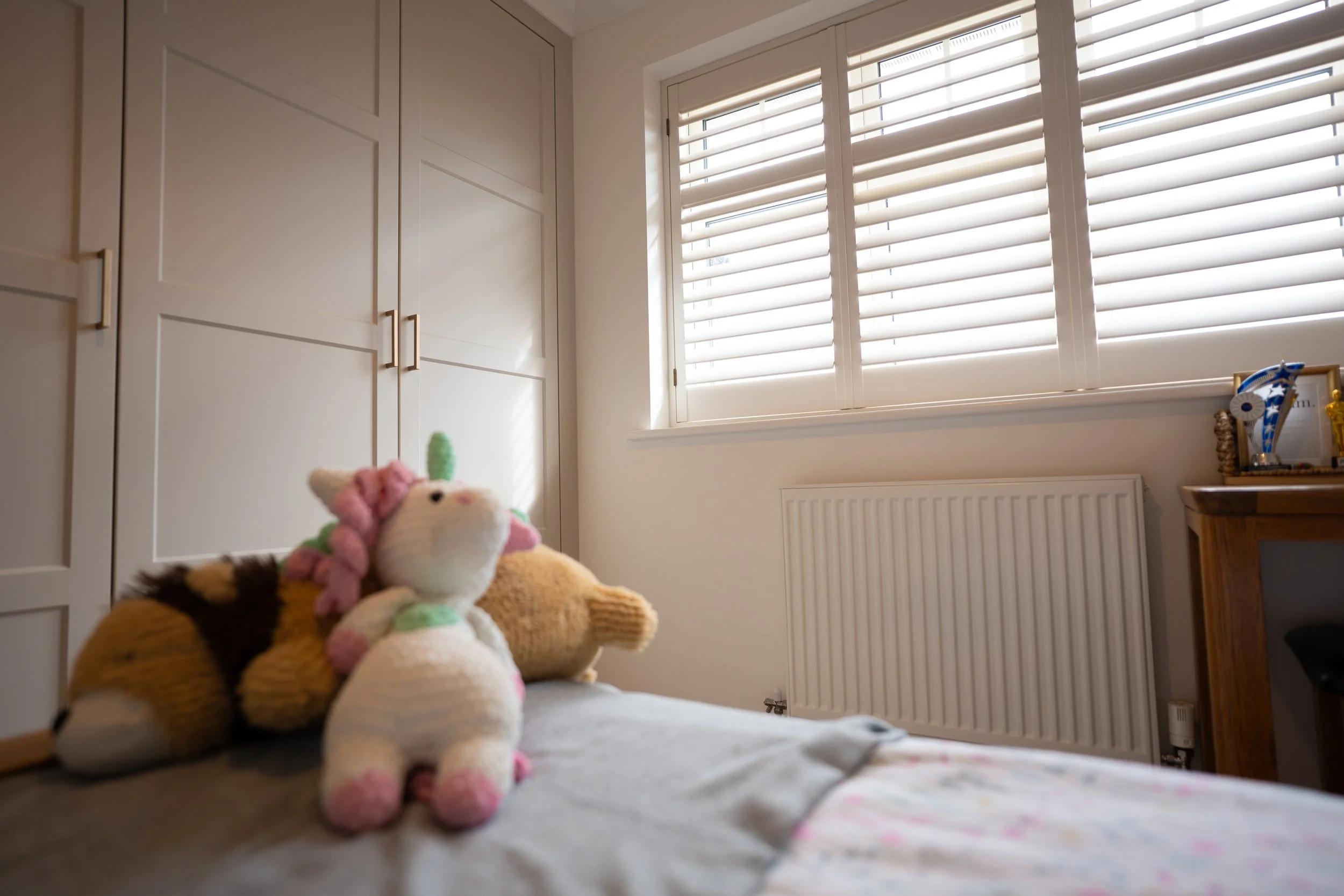 A stuffed animal toy sitting on a bed in a bedroom with closed white window shutters, a white cabinet, and a radiator.