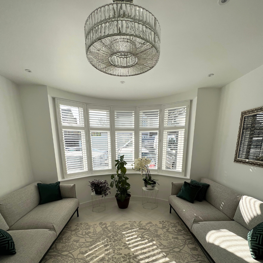 Bright living room with white walls, a large window with white blinds, two beige sofas with dark green pillows, a chandelier, a decorative mirror, a patterned rug, and several potted plants near the window.