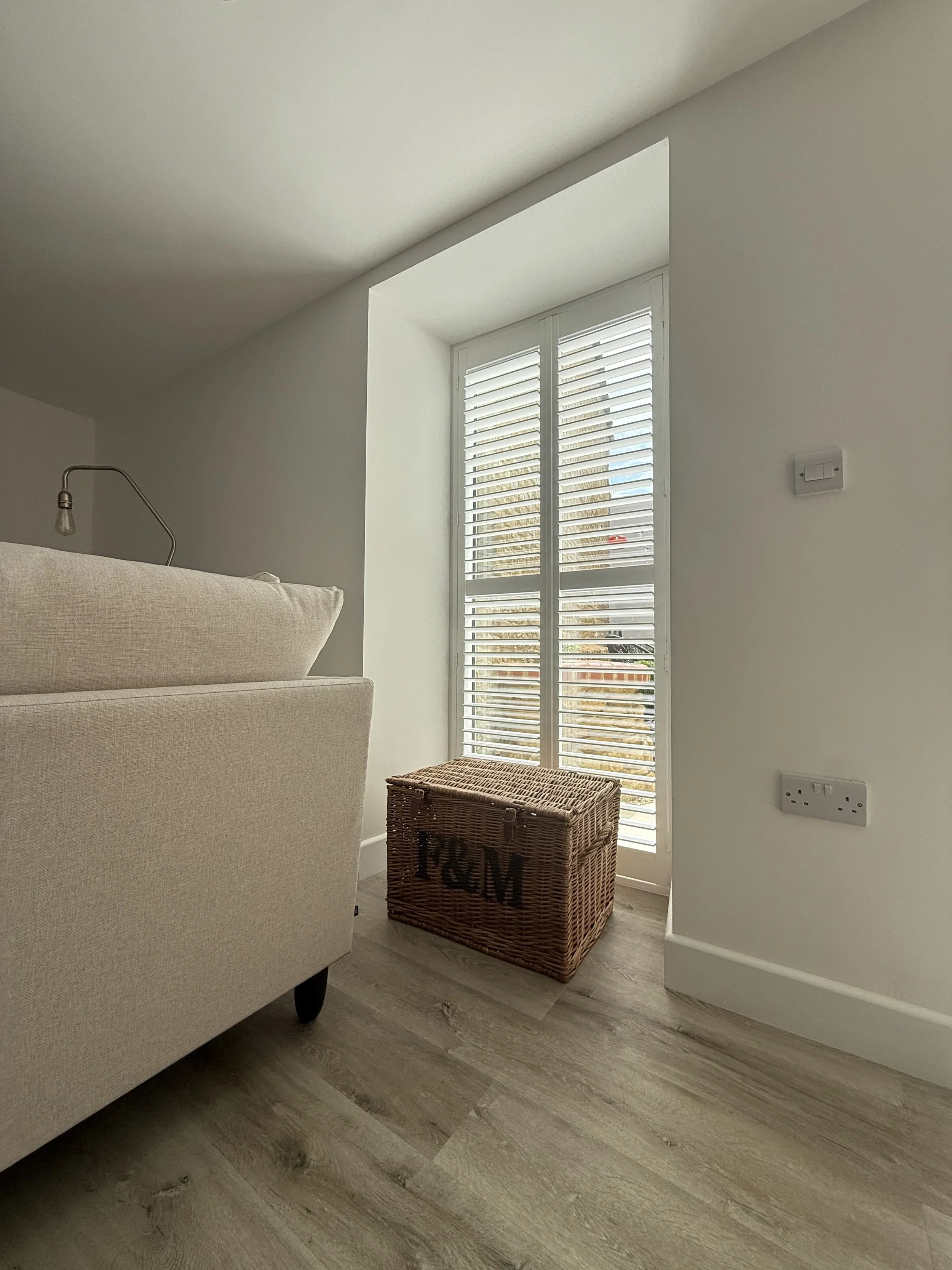Living room with beige sofa, wicker storage box labeled 'FUN,' and window with white shutters letting in light.