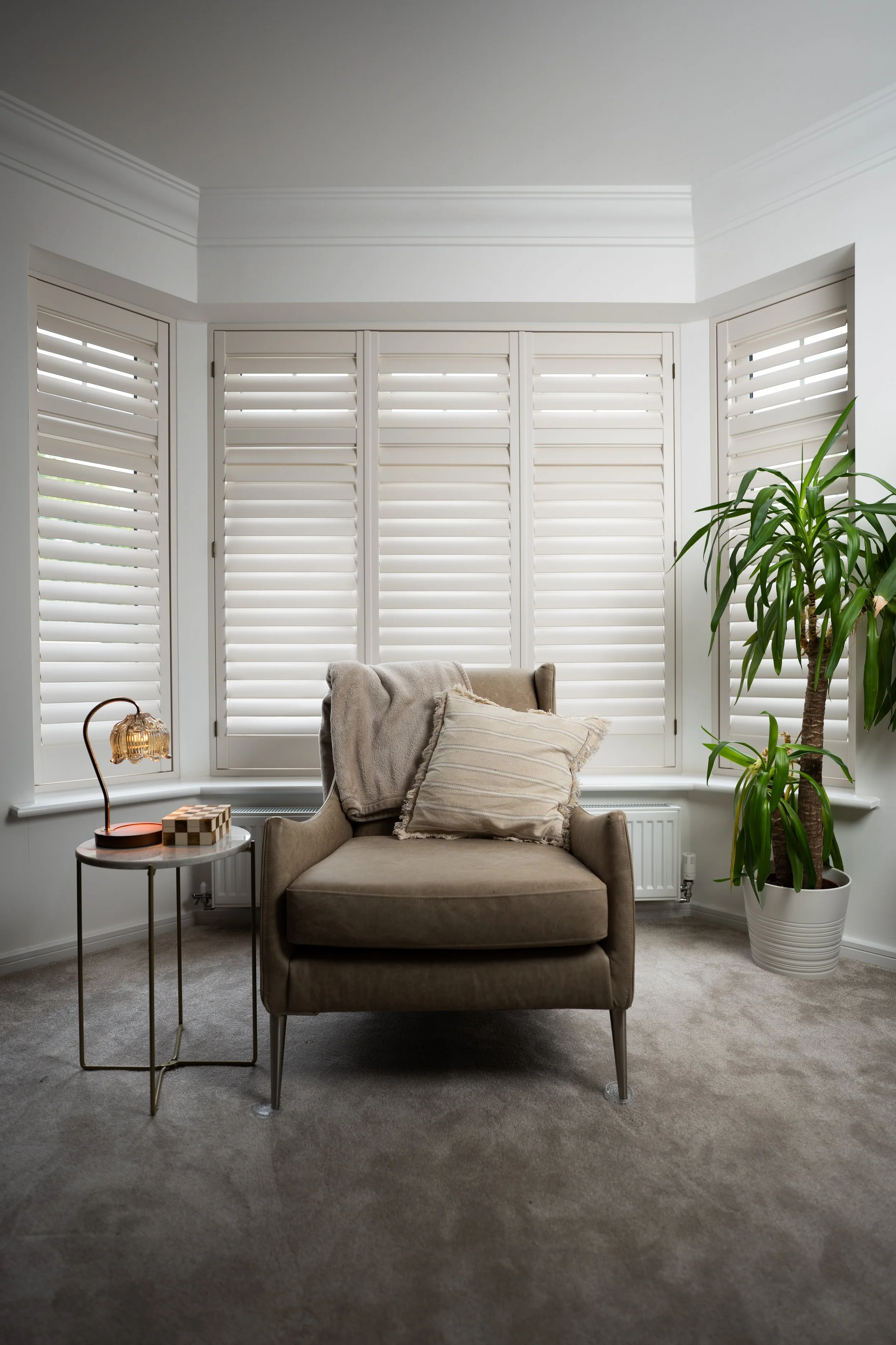 Living room corner with a beige armchair, a side table with a lamp and a decorative box, white plantation shutters on a bay window, a large potted plant, cushions on the armchair, and carpeted flooring.