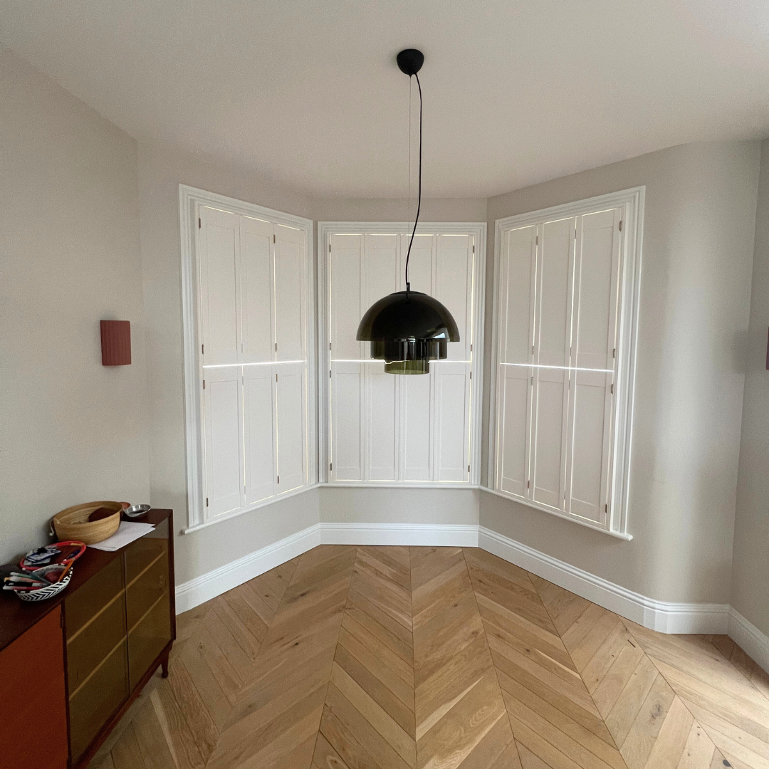Empty room with wooden chevron-patterned floor, white walls, and large windows with white shutters; black pendant light hanging from ceiling.