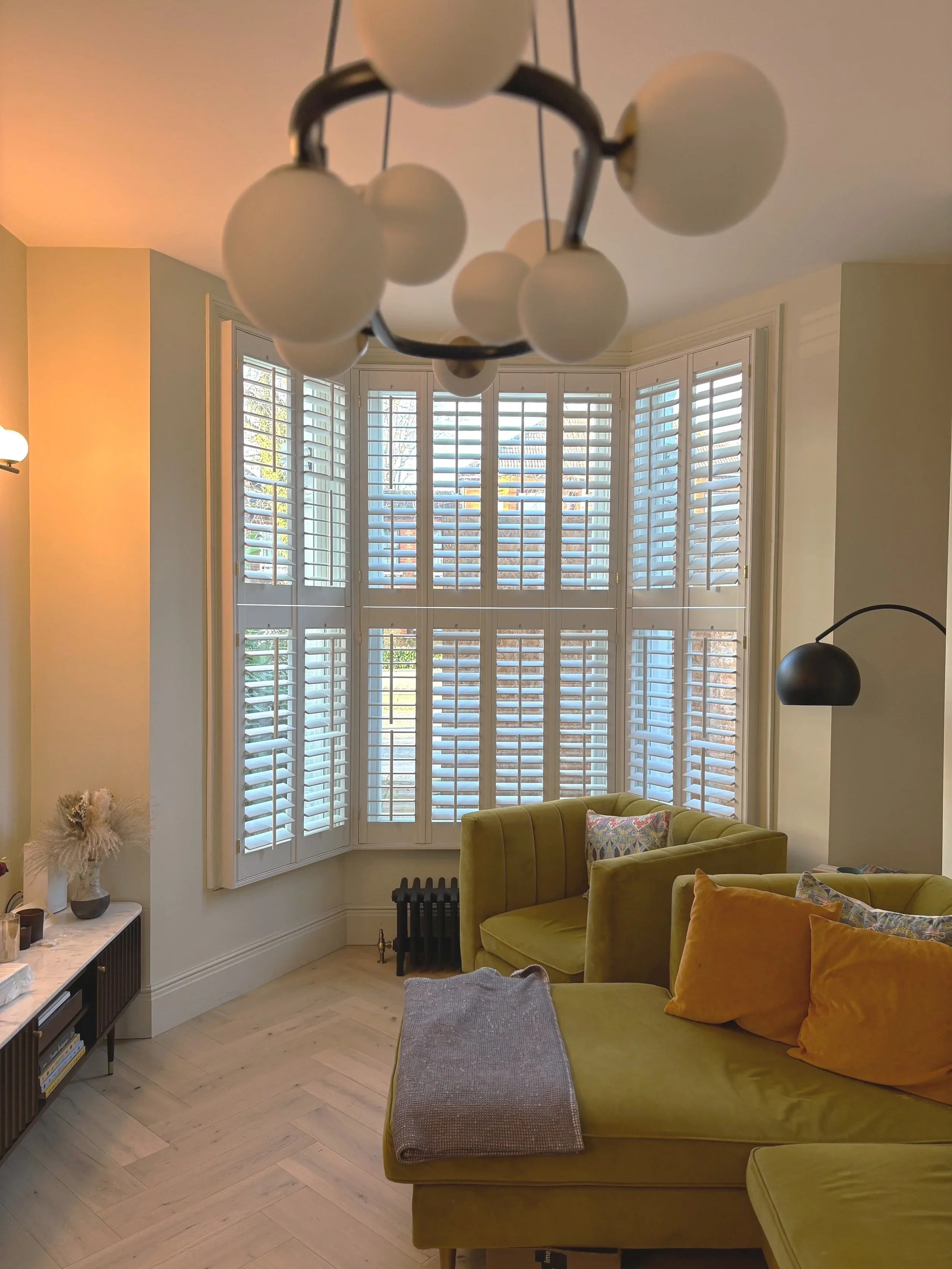 A cozy living room with a rounded yellow-green sectional sofa with orange and patterned pillows. Large bay windows with white plantation shutters let in natural light. A small black radiator is near the window. To the left, a white table holds a vase