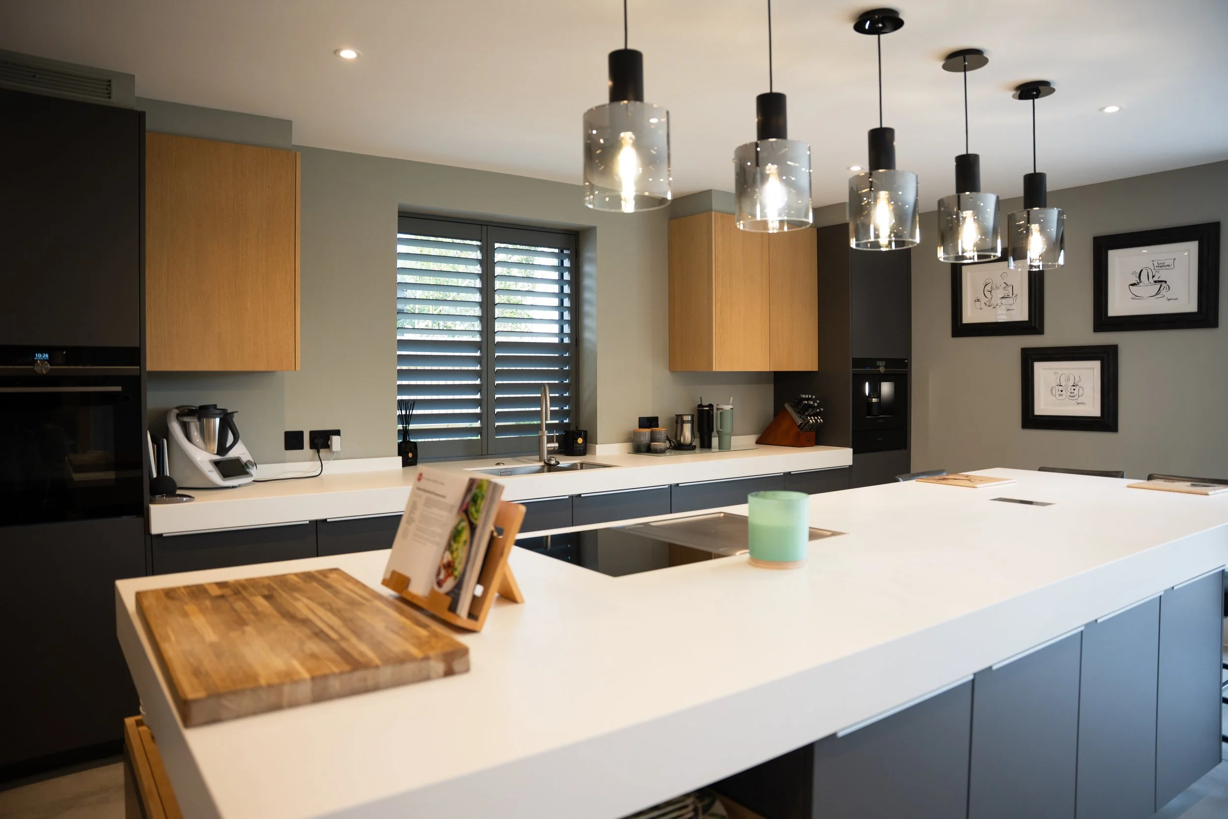 Modern kitchen with pendant lights, a white island countertop, wooden cabinets, framed kitchen-themed artwork, and a window with shutters.