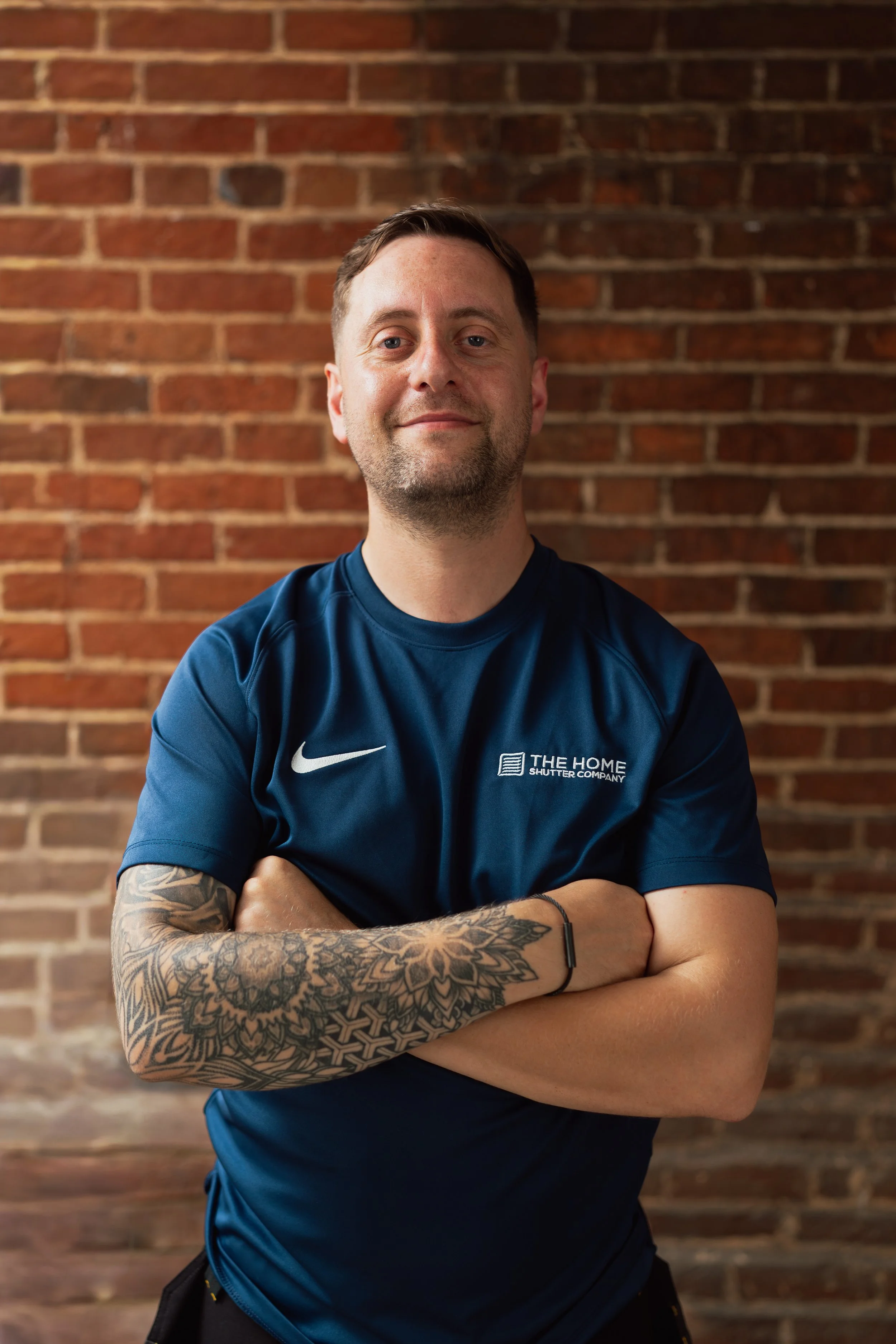 A man with short brown hair, light skin, and tattoos on his right arm, smiling with crossed arms in front of a brick wall. He is wearing a navy blue sports shirt with logos and text, including Nike and 'THE HOME'.