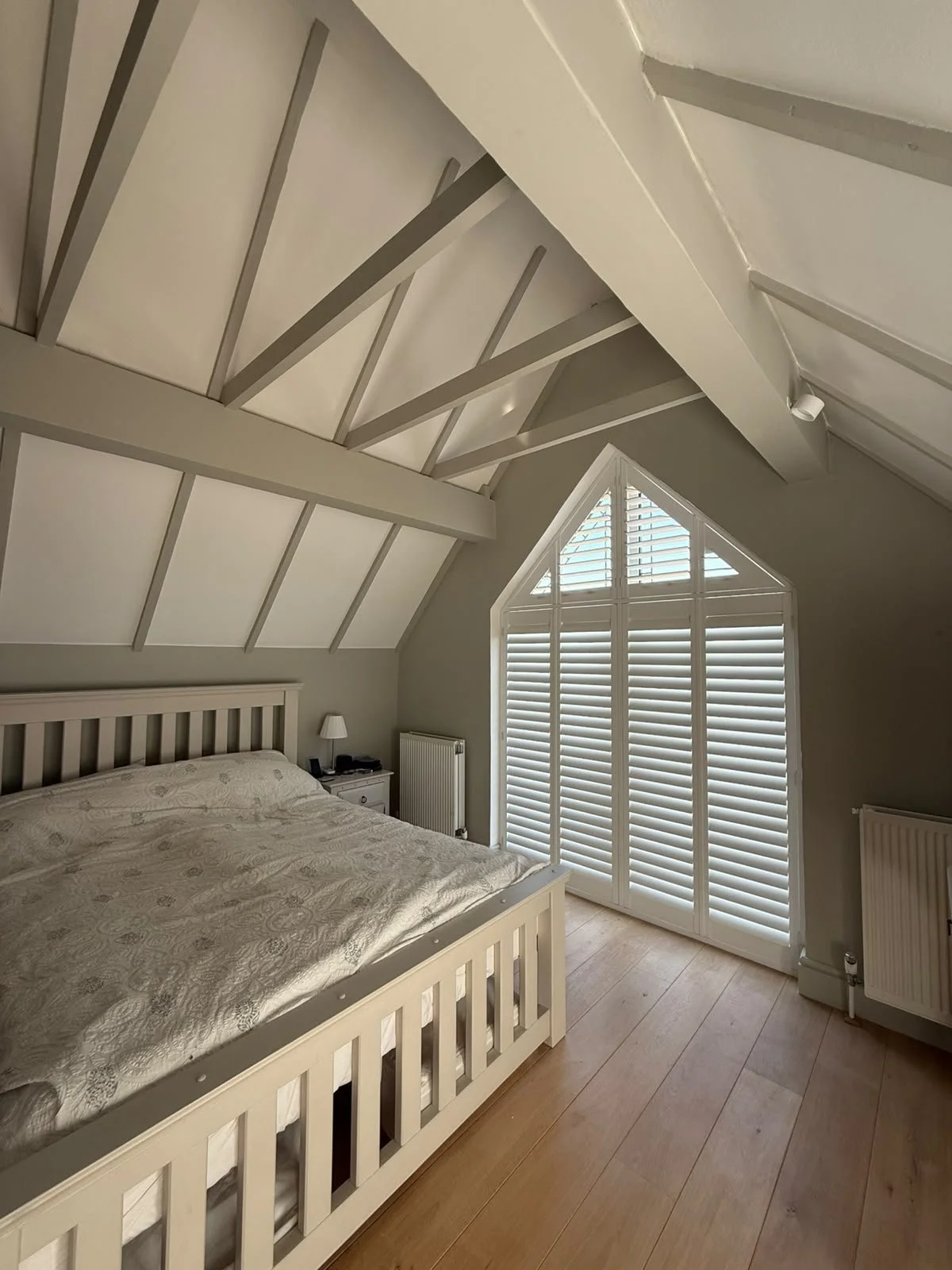 A bedroom with a sloped ceiling, a large window with shutters, a bed with patterned bedding, a nightstand with a lamp, and radiators.