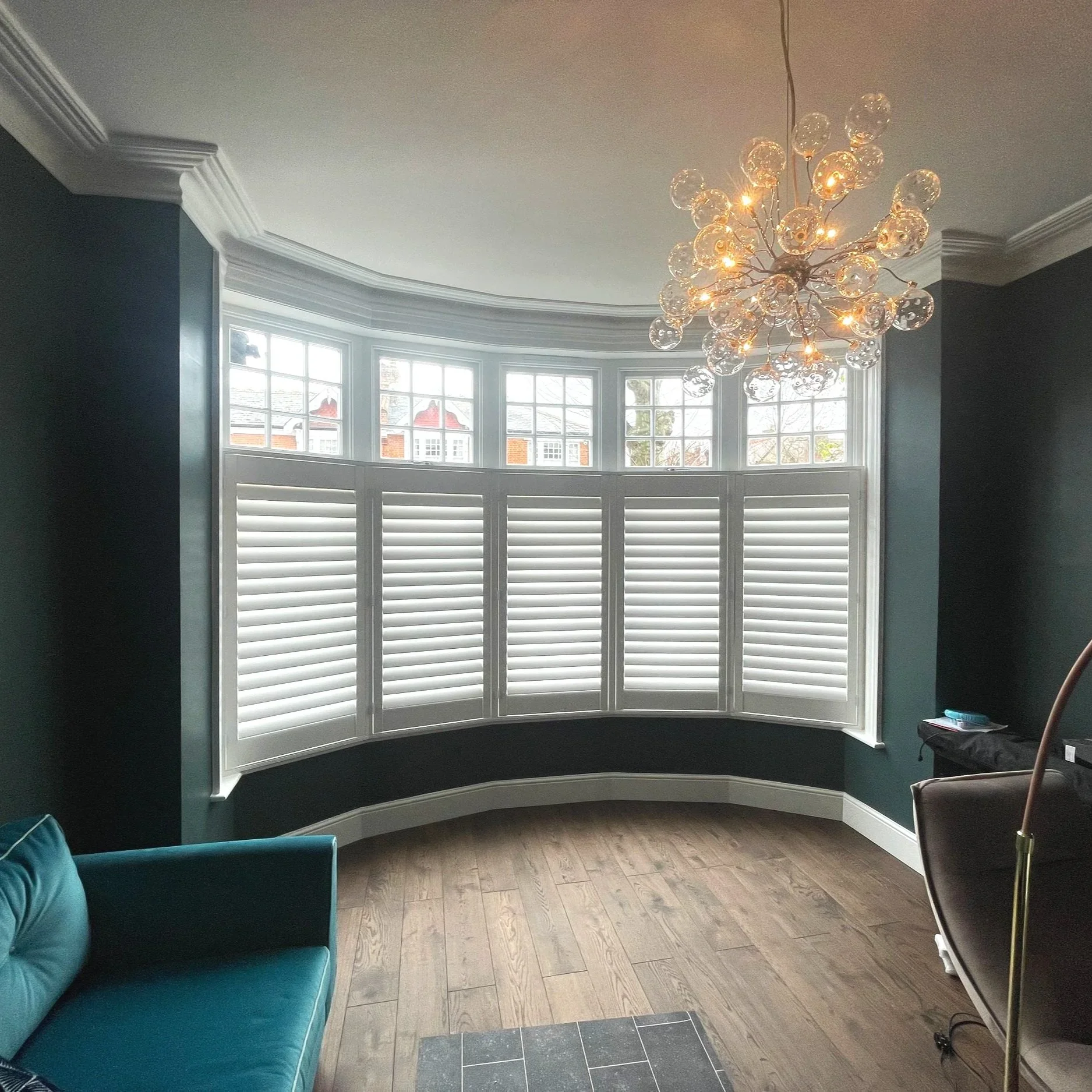 A cozy living room with a large bay window, white shutters, a teal sofa, and a modern chandelier hanging from the ceiling, with hardwood flooring.