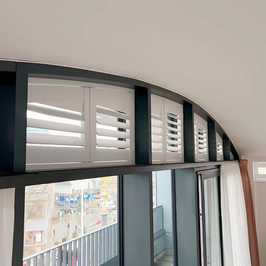 Interior view of a room with a sloped ceiling, blue and white shutters covering upper windows, sliding glass door, and a balcony outside with a city view.