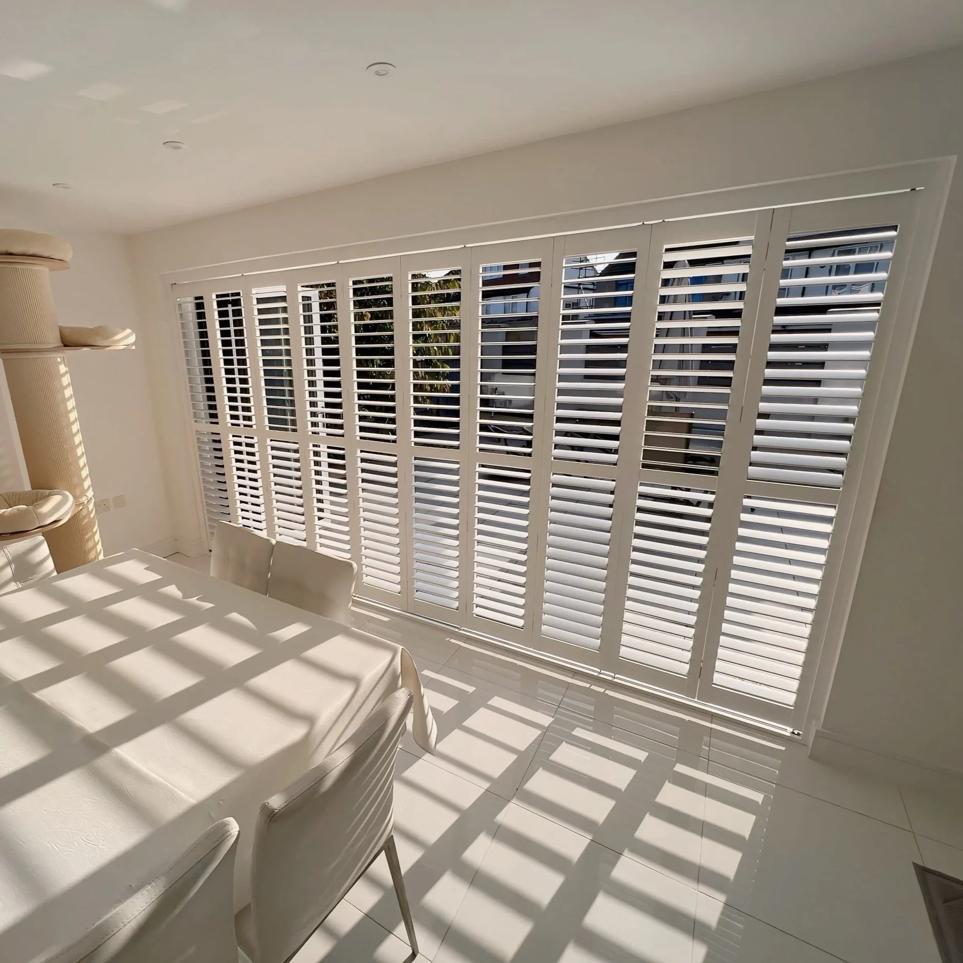 Interior of a room with white shutters on windows, casting striped shadows on the white table and chairs below, with a beige cat tree in the corner.