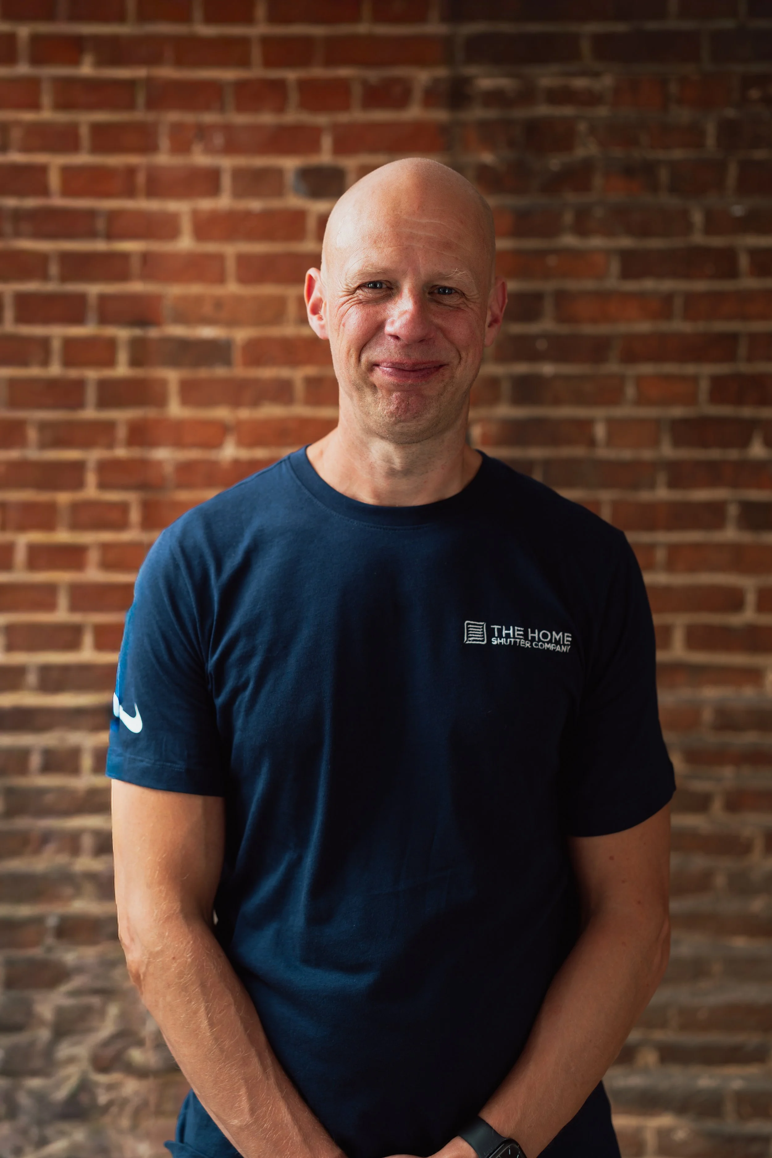 A smiling man with a shaved head wearing a dark blue T-shirt standing against a brick wall.