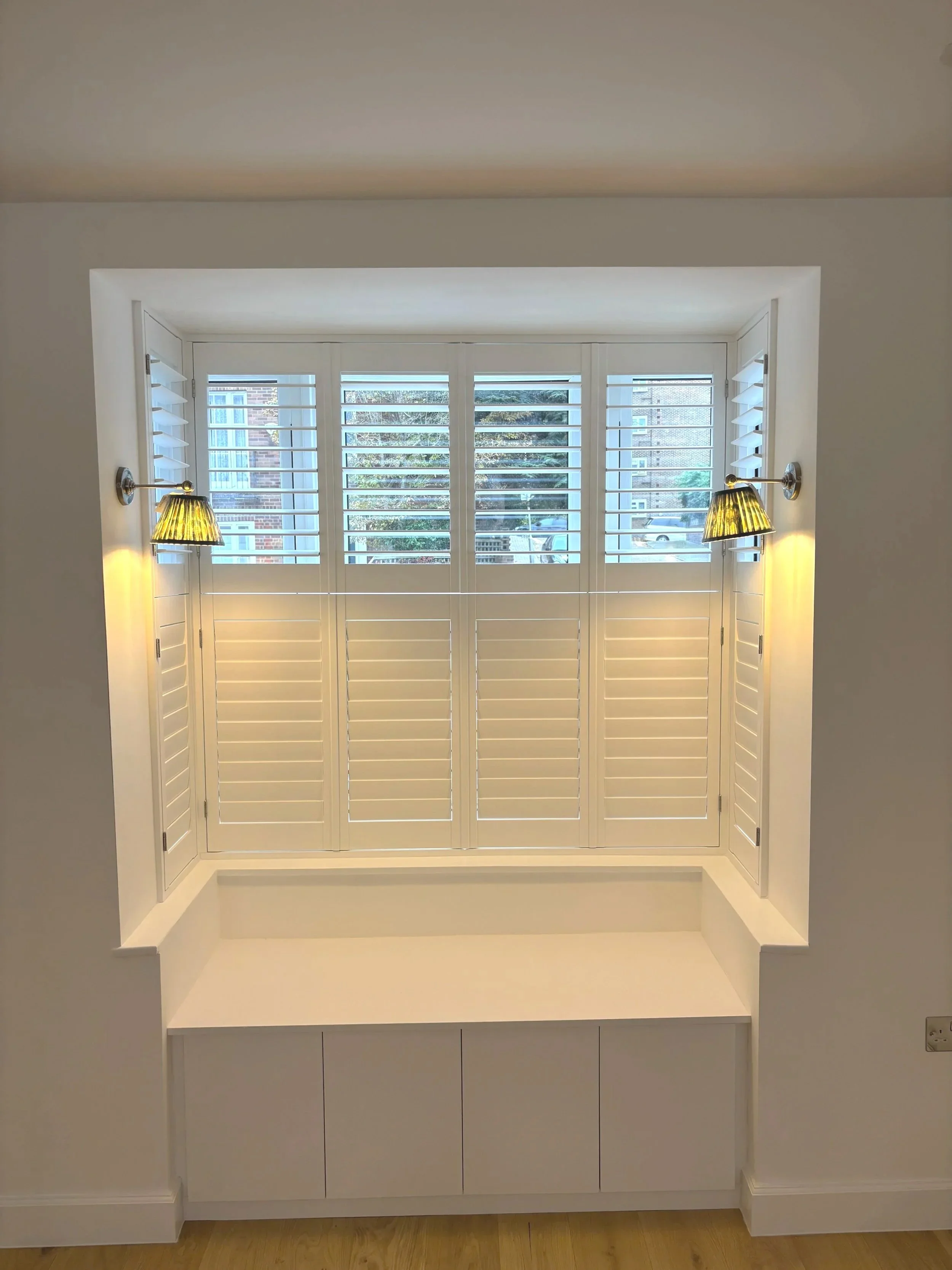 A display window with white plantation shutters, two wall-mounted lamps with yellow shades, and a built-in white cabinet below. Outside the window, a residential street with parked cars and trees is visible.